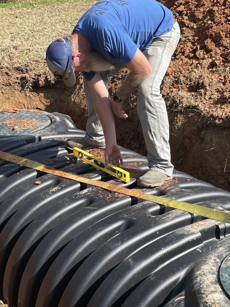 Man in blue shirt levels a black plastic tank in a dirt trench, outdoors.