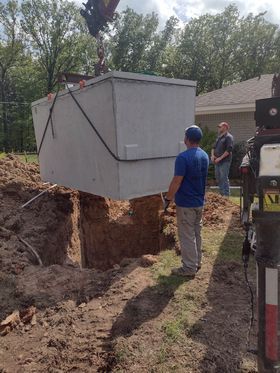 A concrete septic tank is lifted by a crane and lowered into a freshly dug hole. Two men observe in daylight.