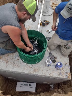 Two men installing equipment into a green basin on a concrete surface, outside. One man wearing a gray shirt and neon green hat.