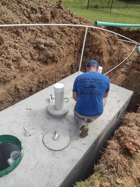 Man in blue shirt kneels beside a concrete septic tank in an open trench, pipes visible.