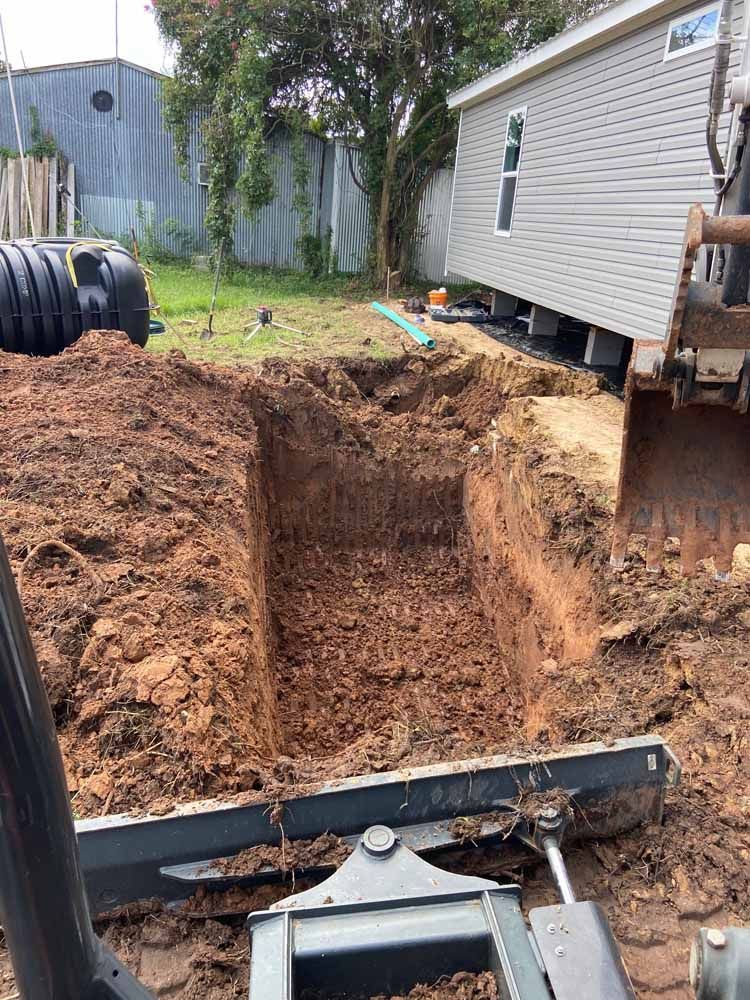 An excavator digging a rectangular pit in a backyard with a house and septic tank in the background.
