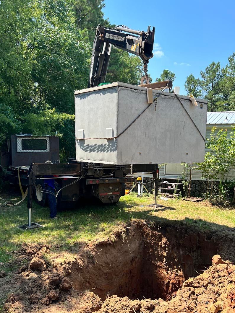 Crane lowering a large concrete septic tank into a dug pit in a backyard setting under a blue sky.