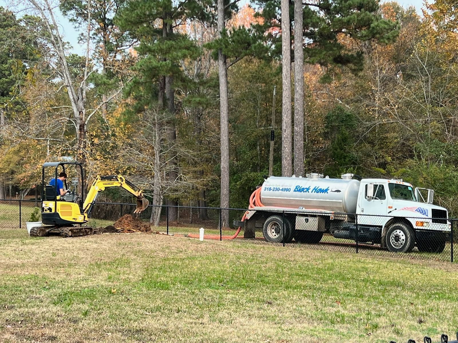 Mini excavator digging near a septic tank truck in a grassy yard, trees in the background.