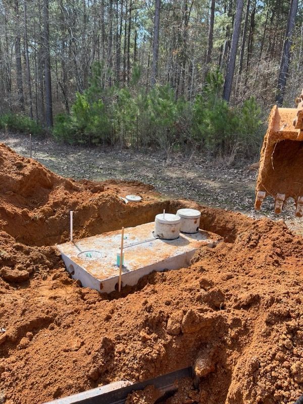 A septic tank in a dirt pit, being worked on by an excavator in a wooded area.