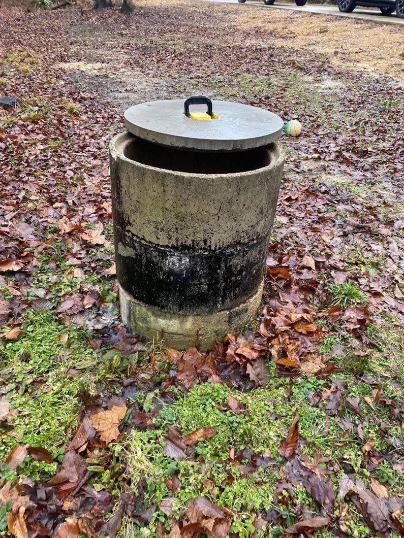Concrete cylinder with a partially open lid sits among leaves and grass.