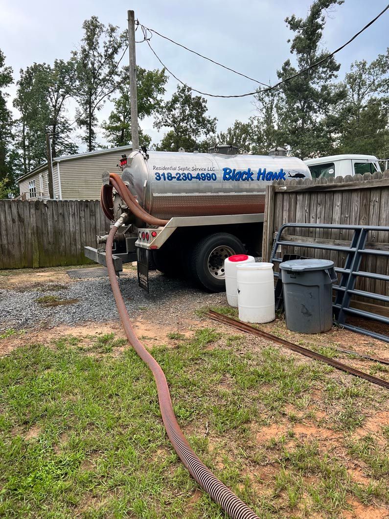 A sewage truck with a hose in a yard. The truck is silver and brown.