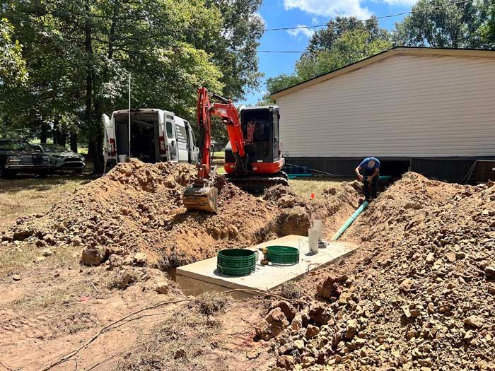 A mini-excavator and workers install a septic tank in a yard near a house.