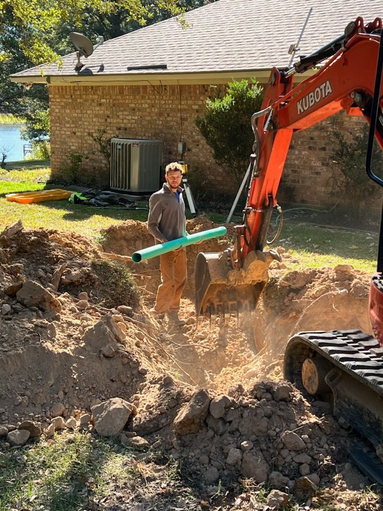 Man in trench holding pipe, next to a Kubota excavator digging. Brick house in background.