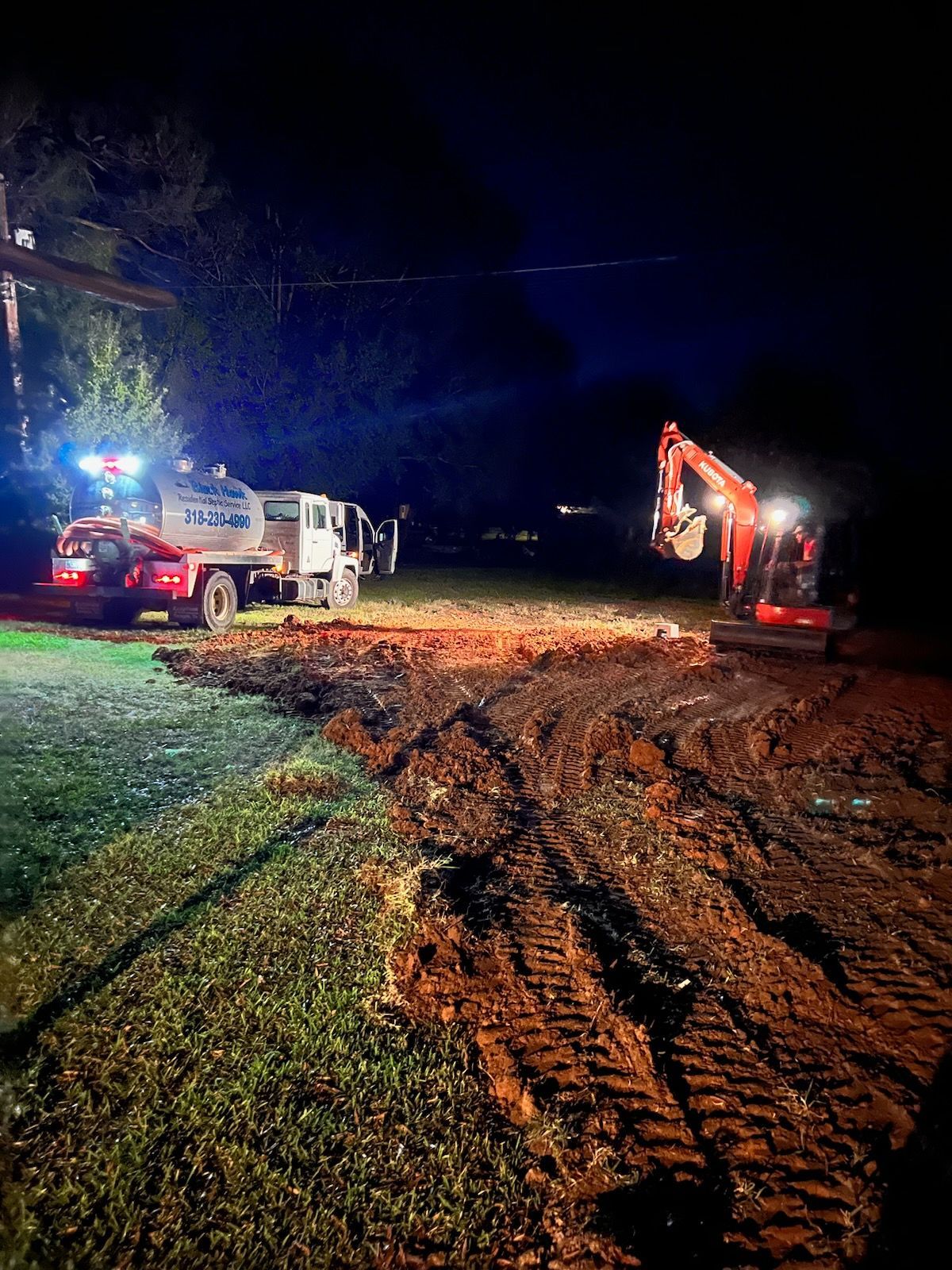 Night construction scene: excavator digging in a muddy field, water truck with lights, power lines overhead.