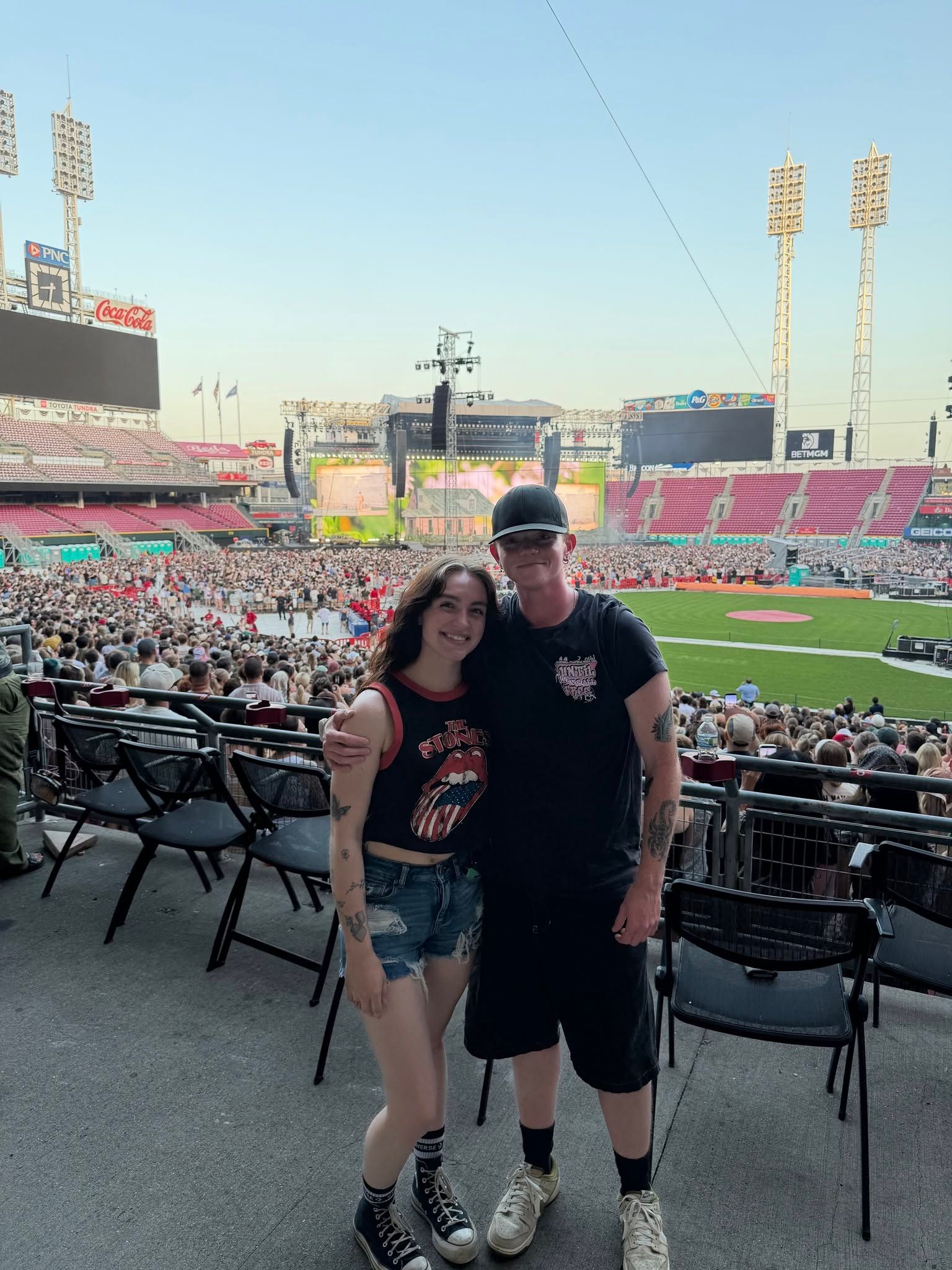 Two fans posing in a stadium with the field and crowd behind them at sunset