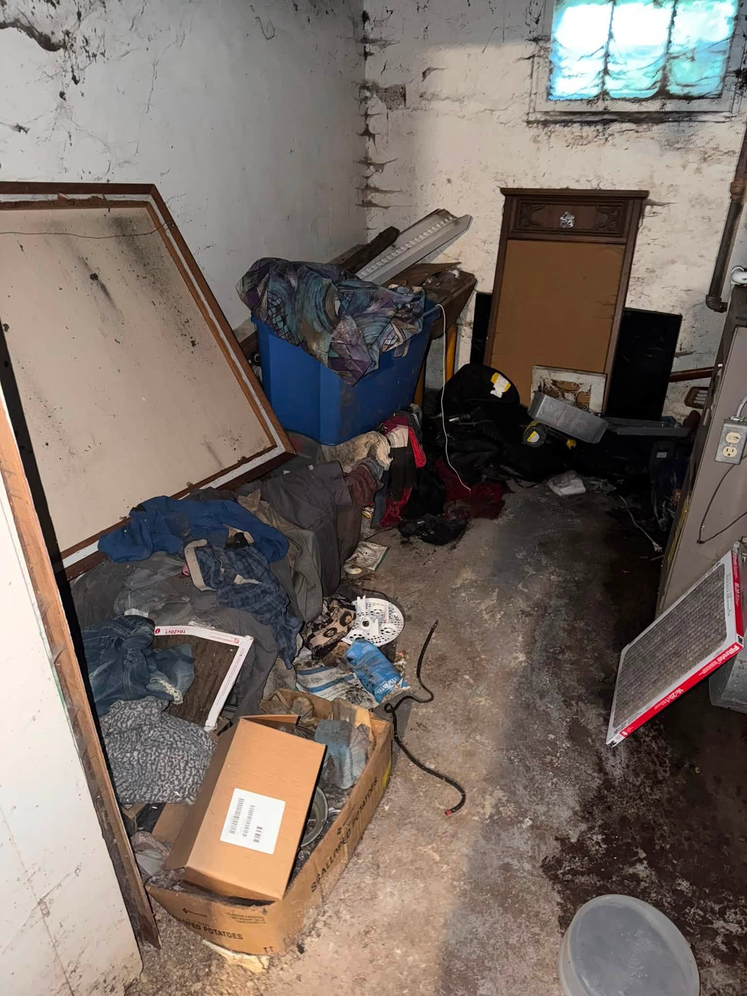Cluttered basement storage room with boxes, bins, scattered items, and a small window high on the wall.