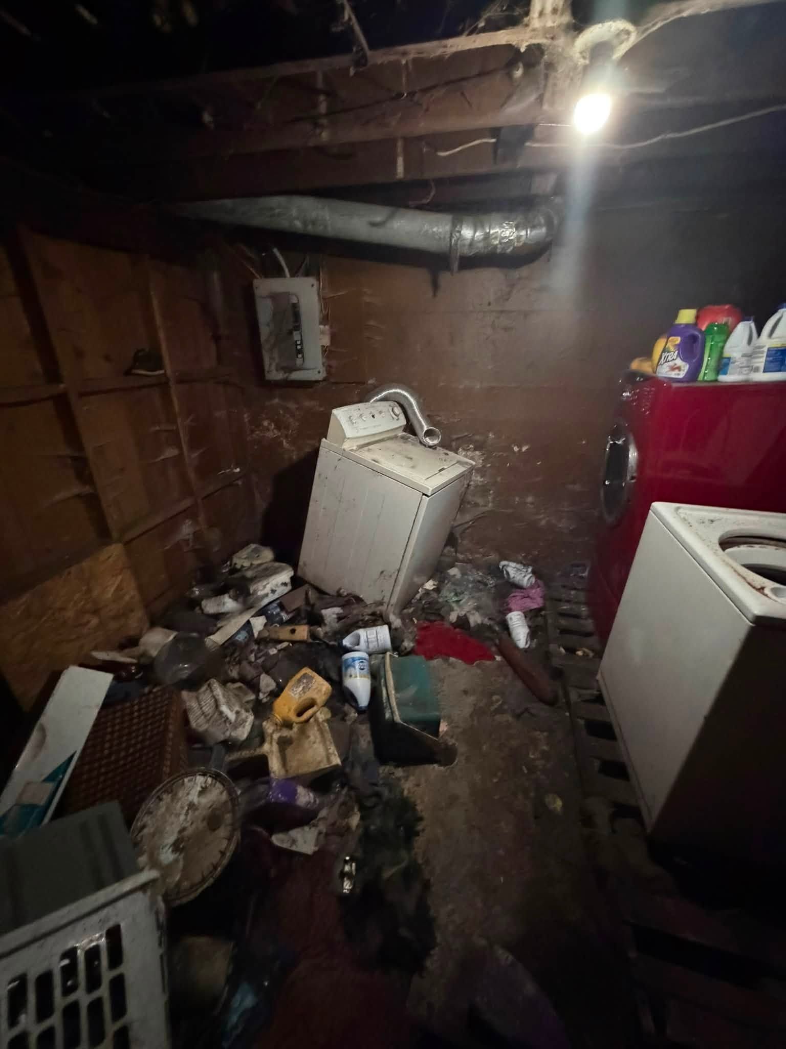 Cluttered basement with debris, tipped white appliance, and exposed pipes under a bare lightbulb.