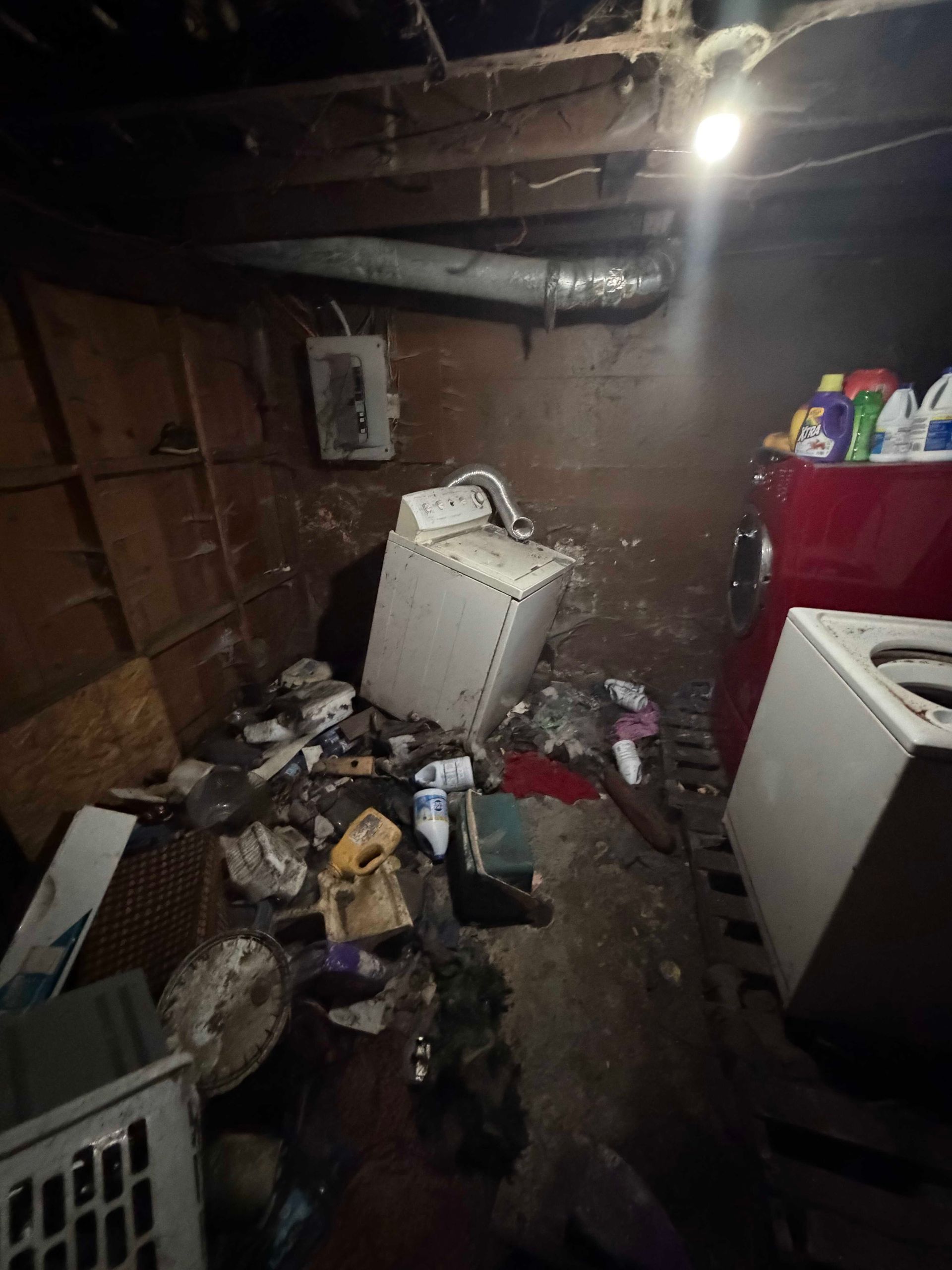 Cluttered basement with trash, a broken appliance, and exposed pipes under a bare light.