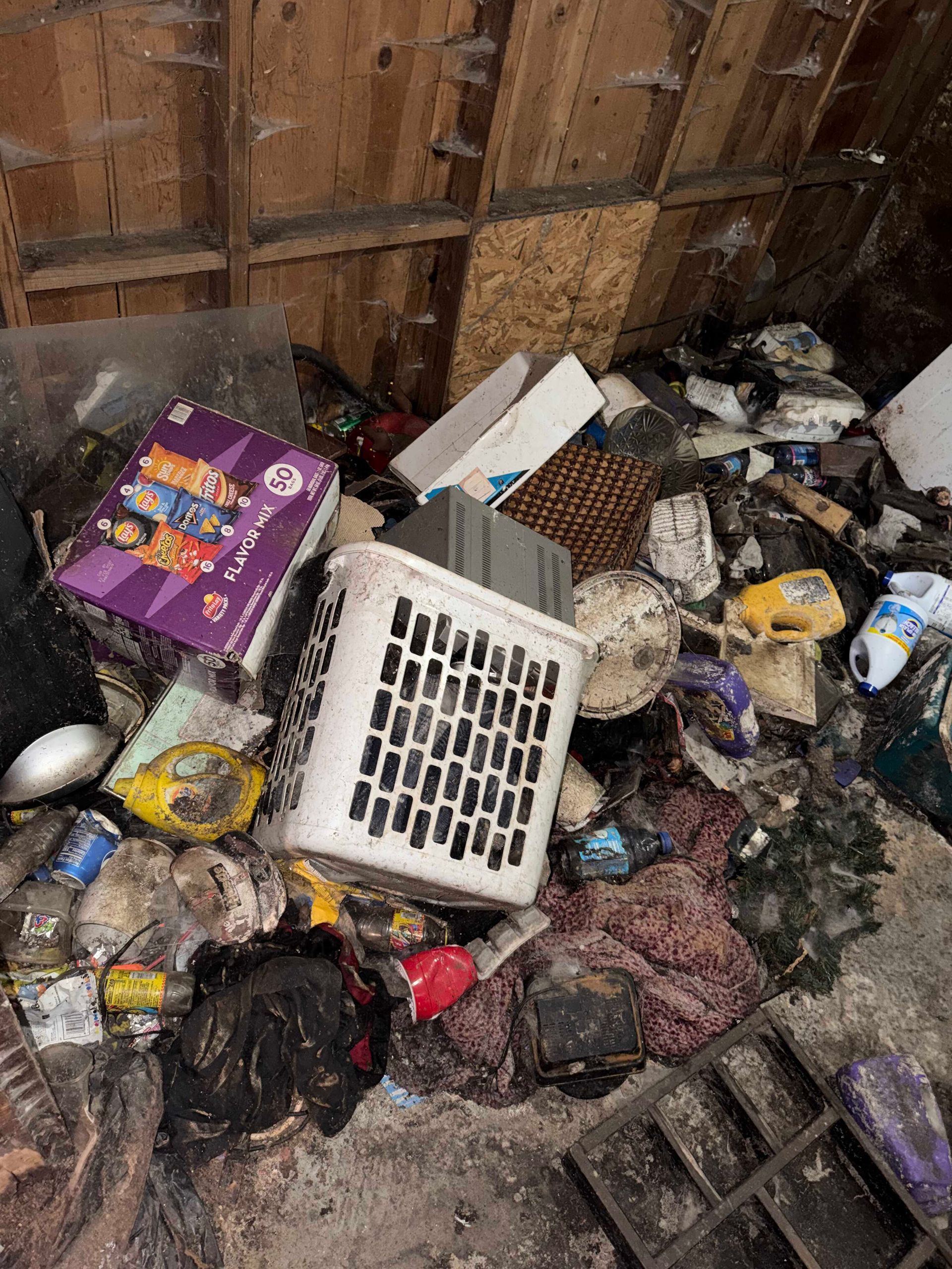 Overflowing trash heap in a wooden enclosure with scattered food containers and debris.