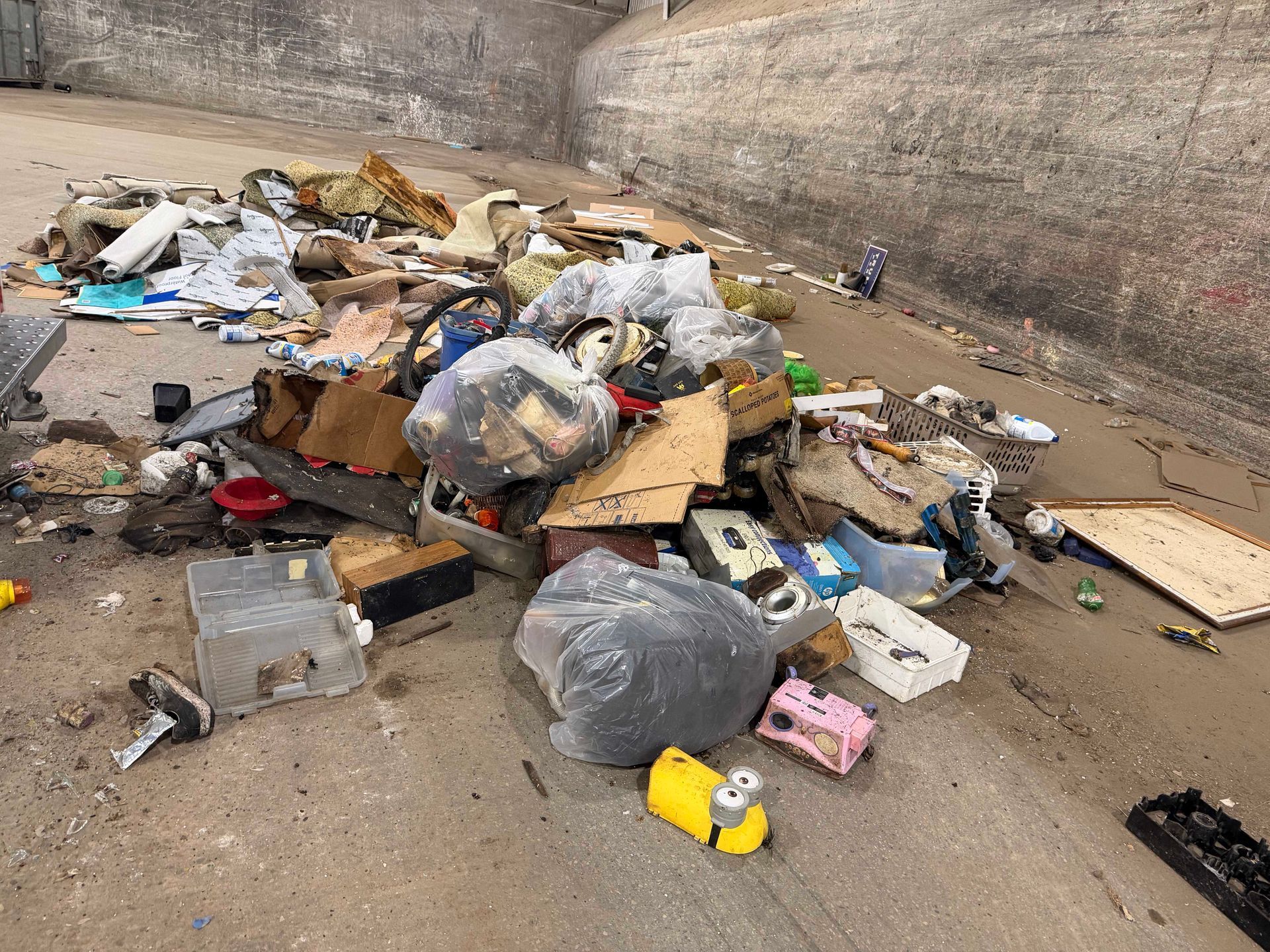 Pile of trash and garbage bags scattered on a concrete floor against a stained wall.