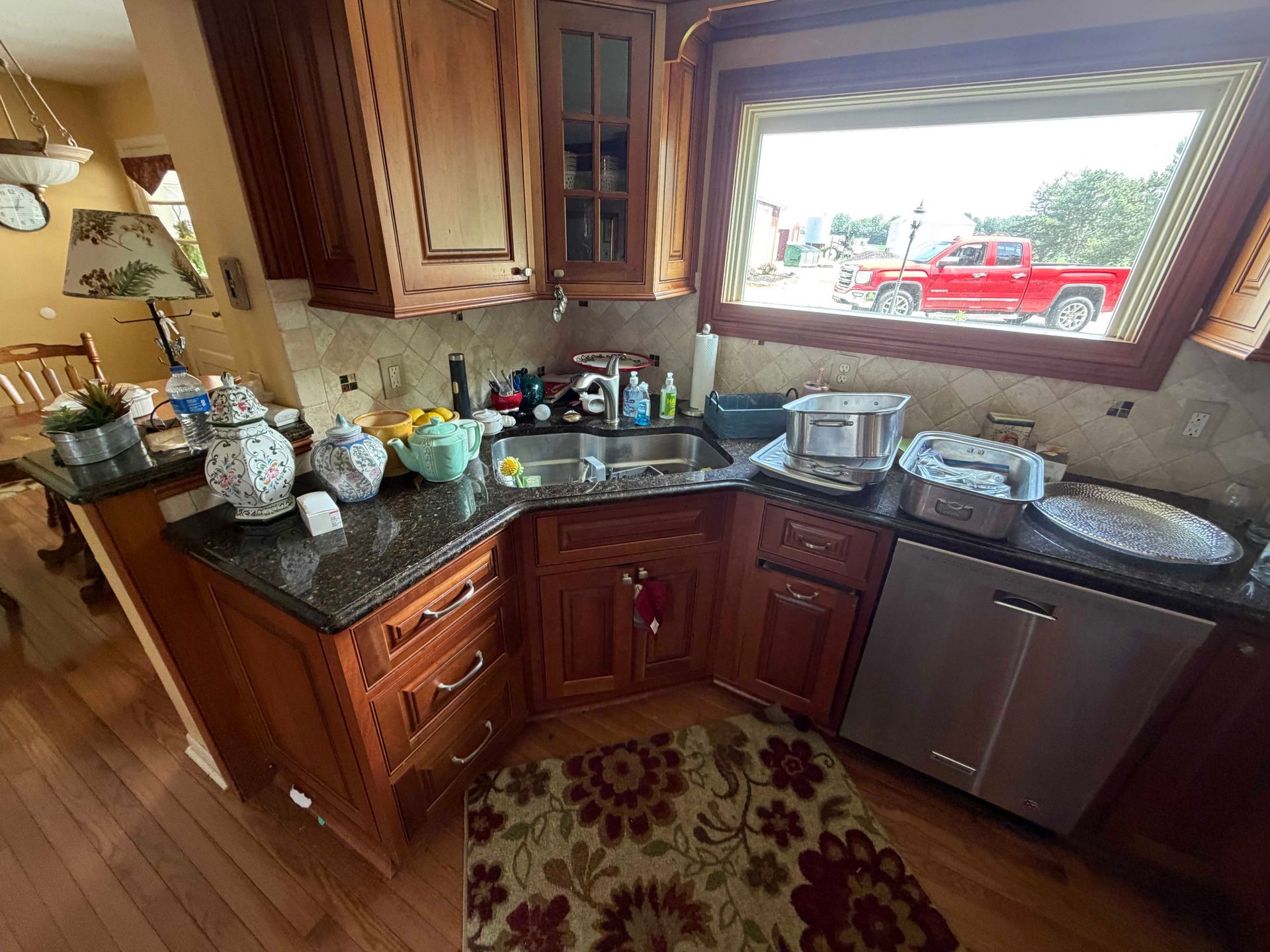 Cluttered kitchen corner with dark wood cabinets, granite counters, dishes, and a window above the sink