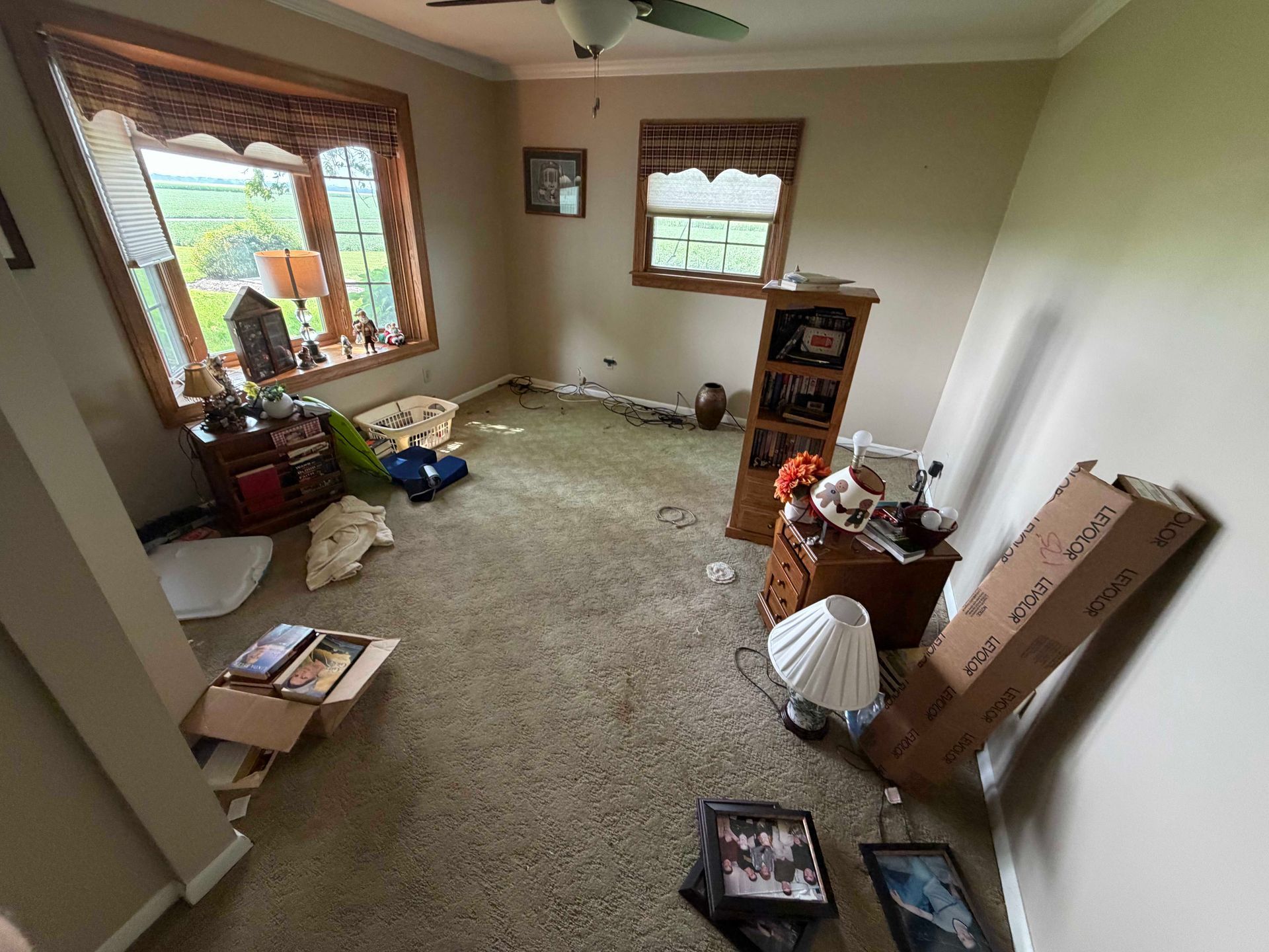 Cluttered beige room with two windows, boxes, and a small shelf on carpeted floor