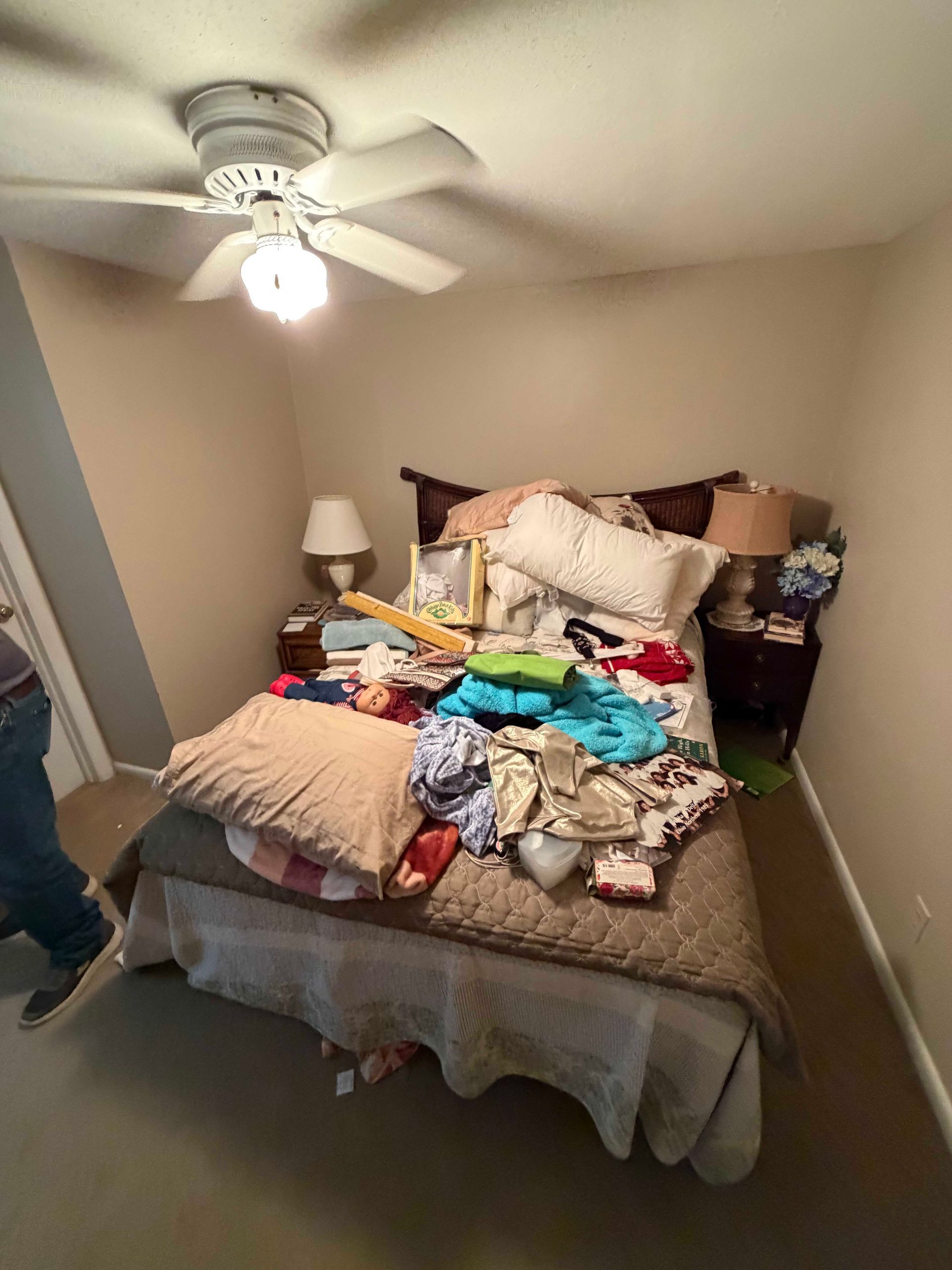 Cluttered bedroom with an unmade bed covered in clothes and pillows, ceiling fan above, lamp on bedside table.