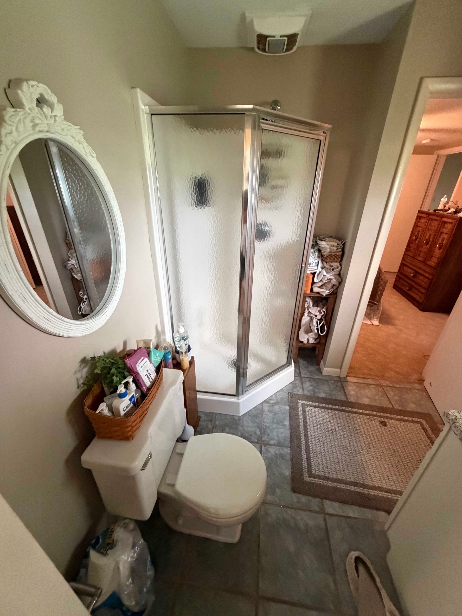 Bathroom with shower stall, toilet, sink, mirror, and beige tile floor leading to a hallway.