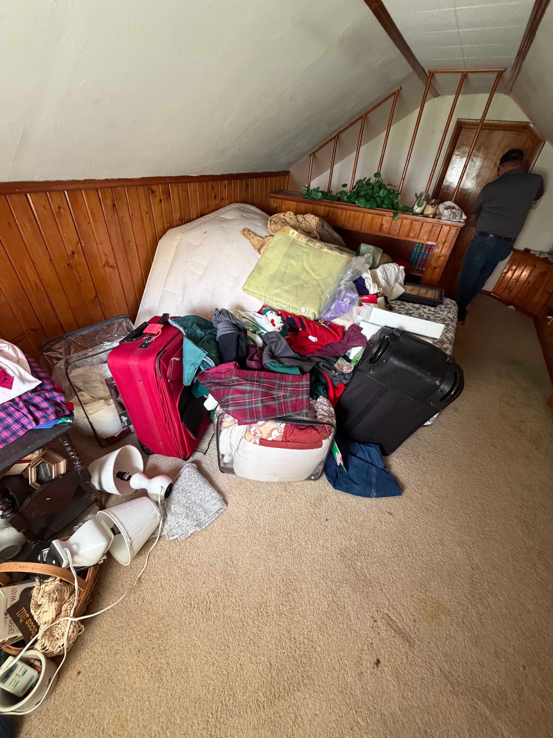 Cluttered attic room with piles of luggage, clothes, and storage bins under a sloped ceiling.