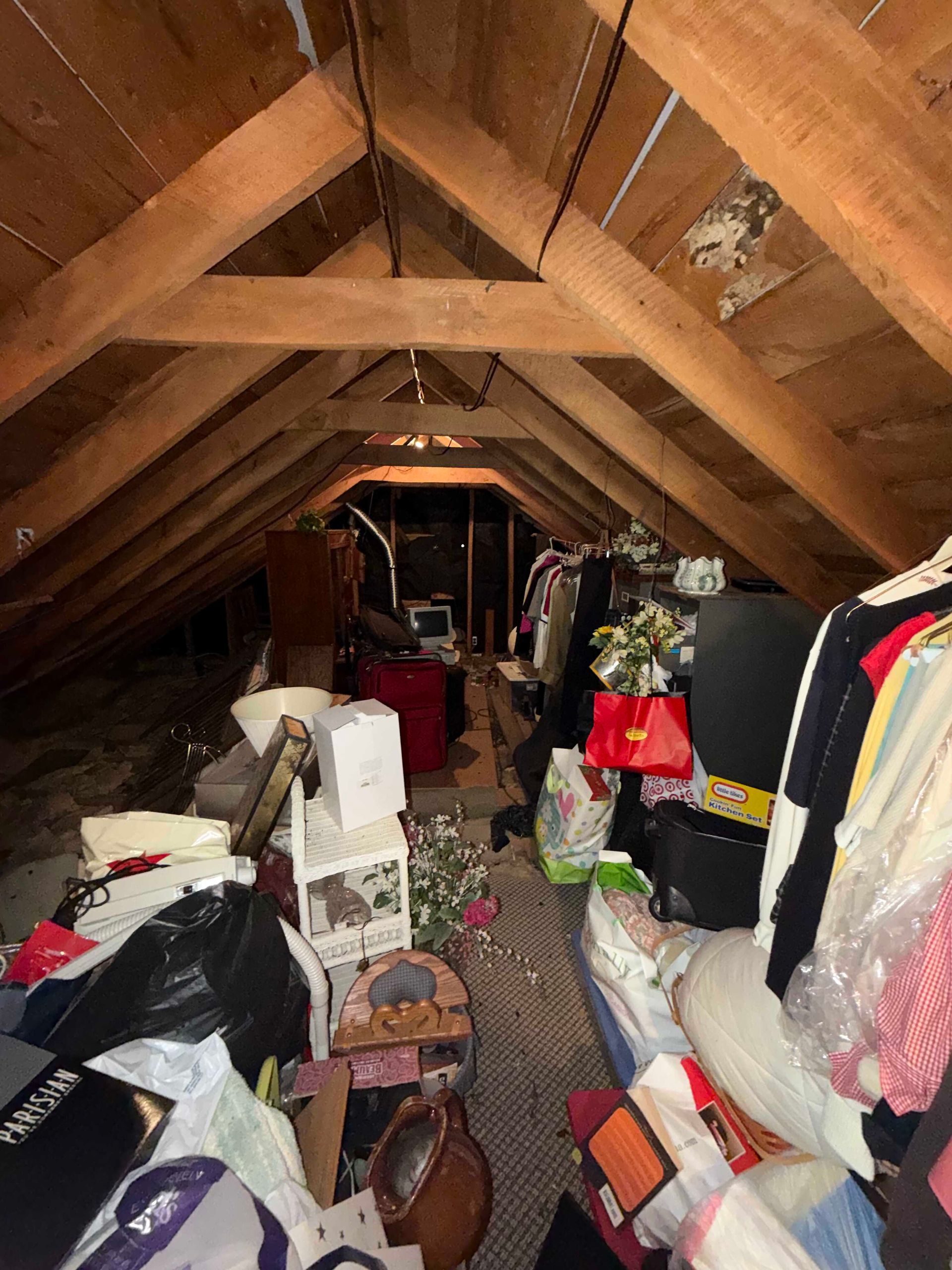 Cluttered attic with boxes, bags, clothes, and assorted stored items under a sloped wooden roof.