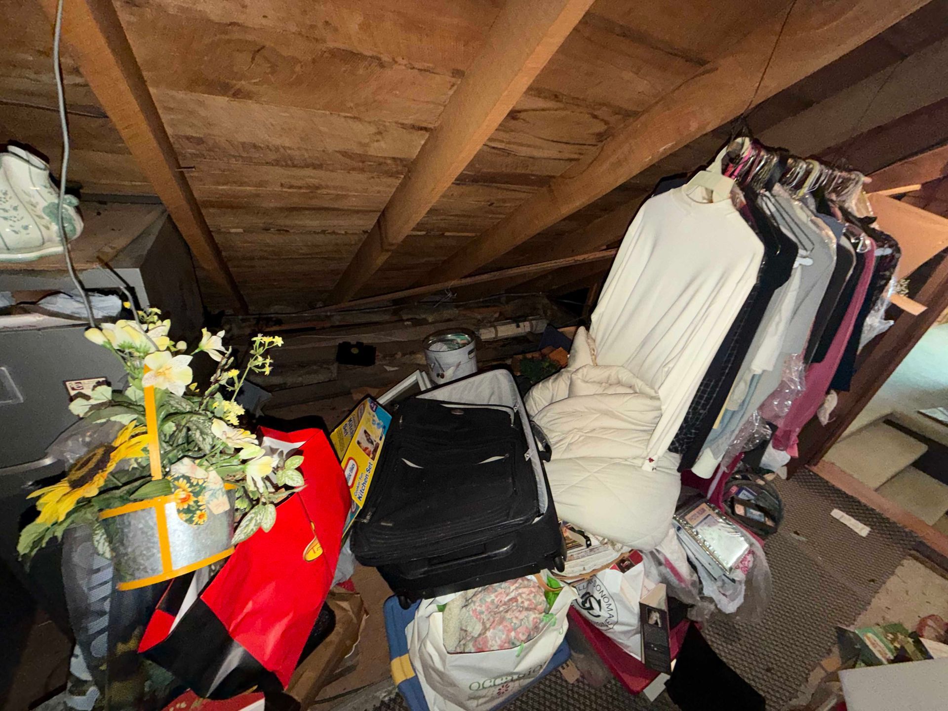 Cluttered attic storage with stacked bags, folded clothes, and a decorated plant under wooden rafters
