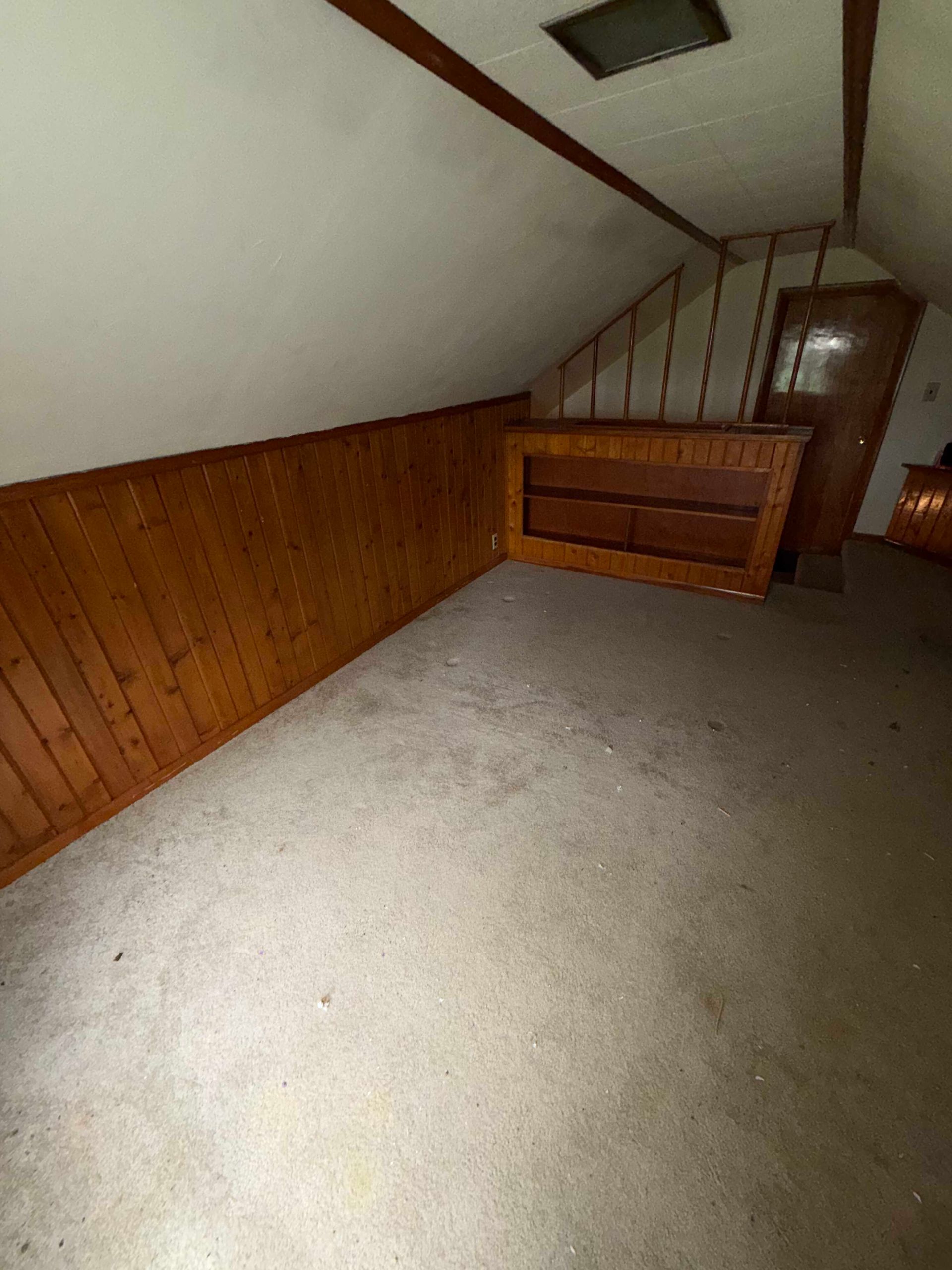 Empty attic room with sloped ceiling, wood paneling, carpet, and a wooden dresser against the wall.