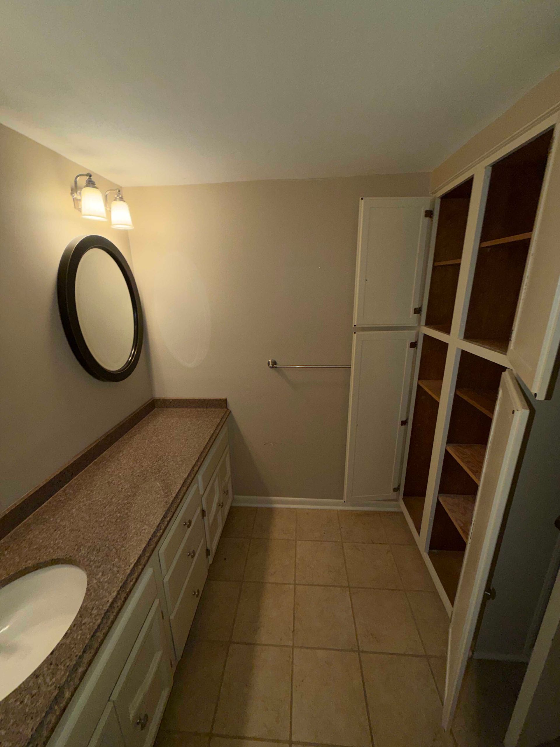 Bathroom with long vanity, round mirror, wall light, beige tile floor, and open shelving cabinet.