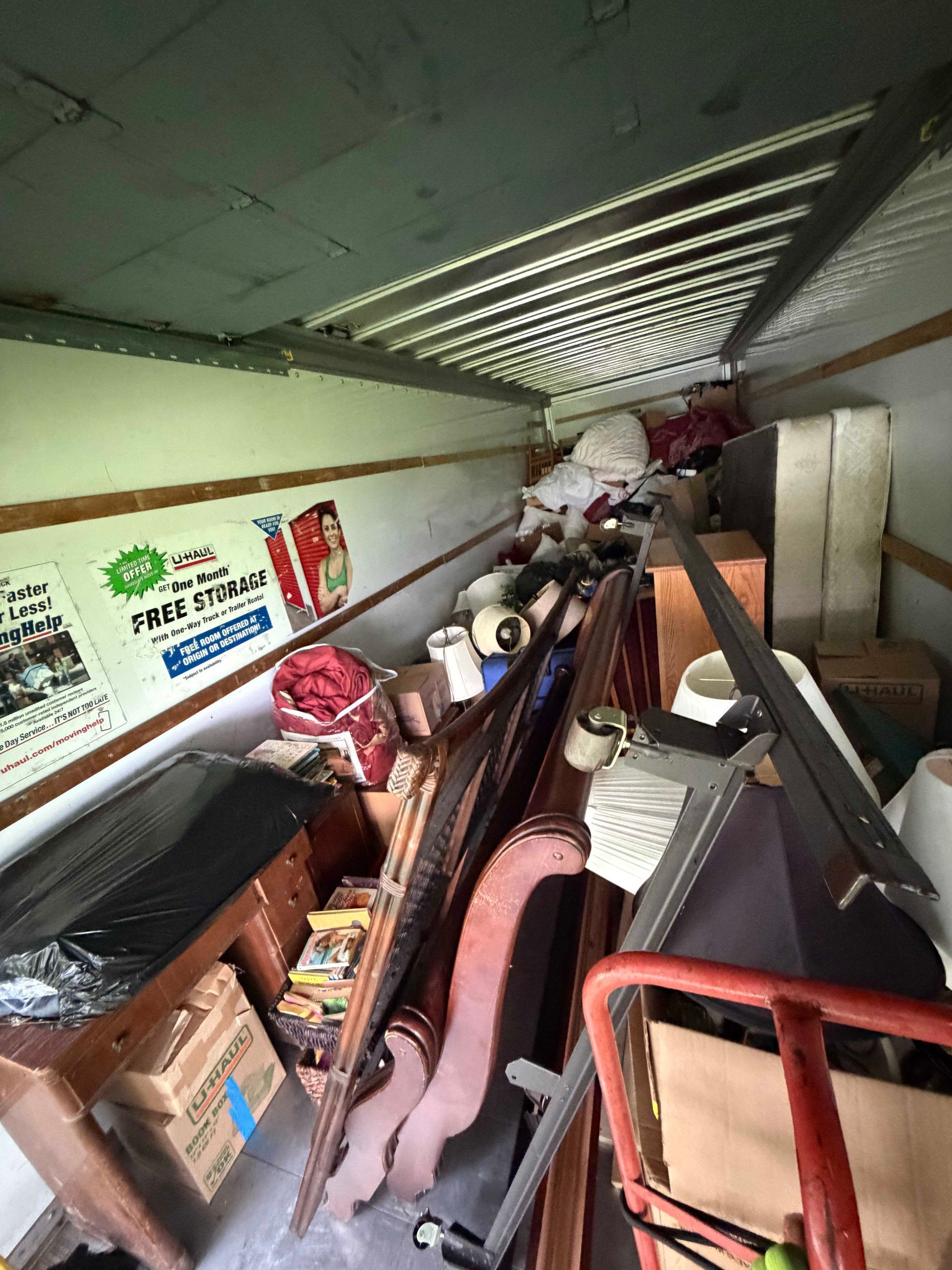 Overcrowded storage room with stacked furniture, boxes, and assorted items along a narrow aisle.