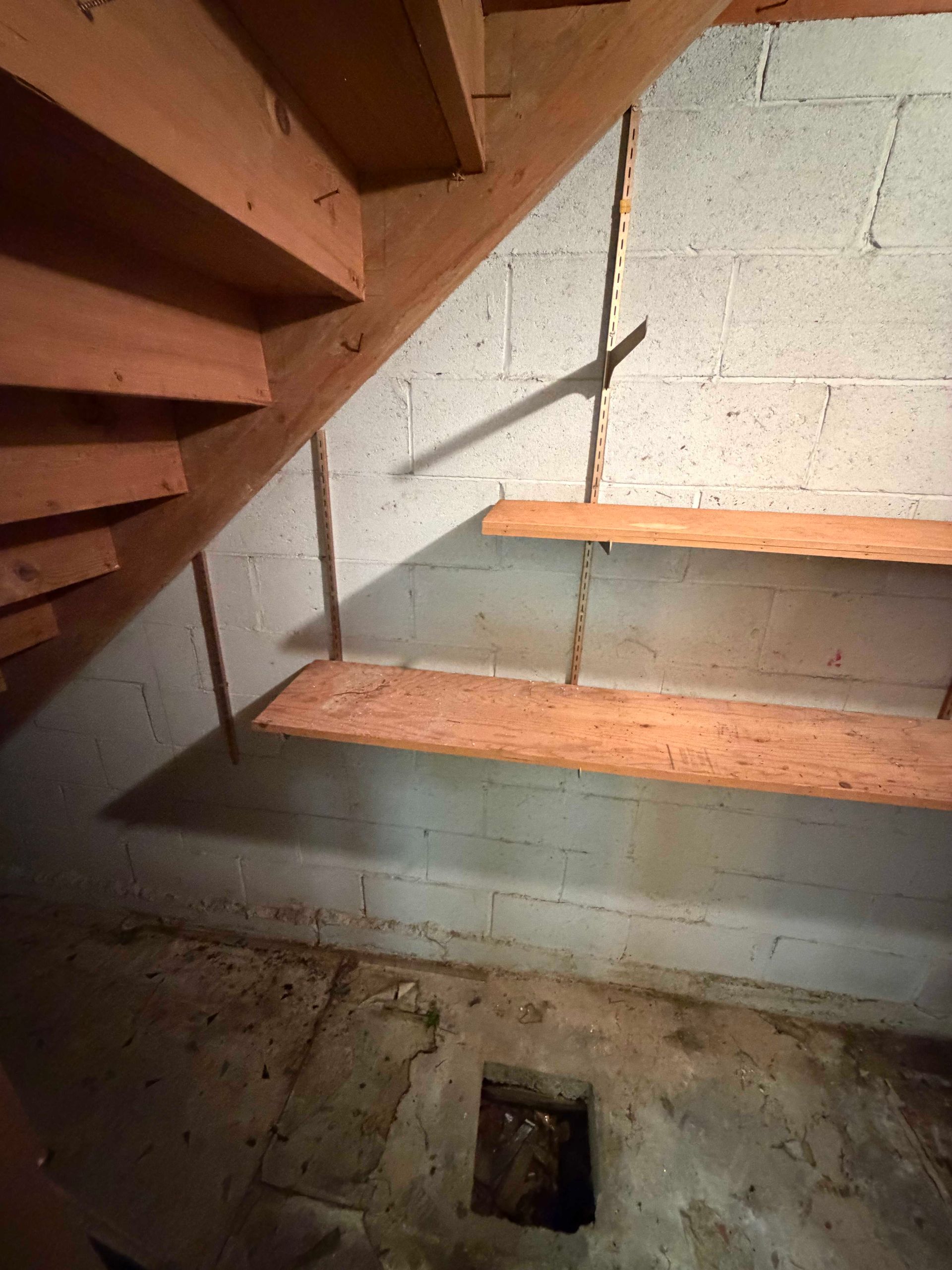 Wooden staircase in a basement, with concrete block wall and a small floor opening below.