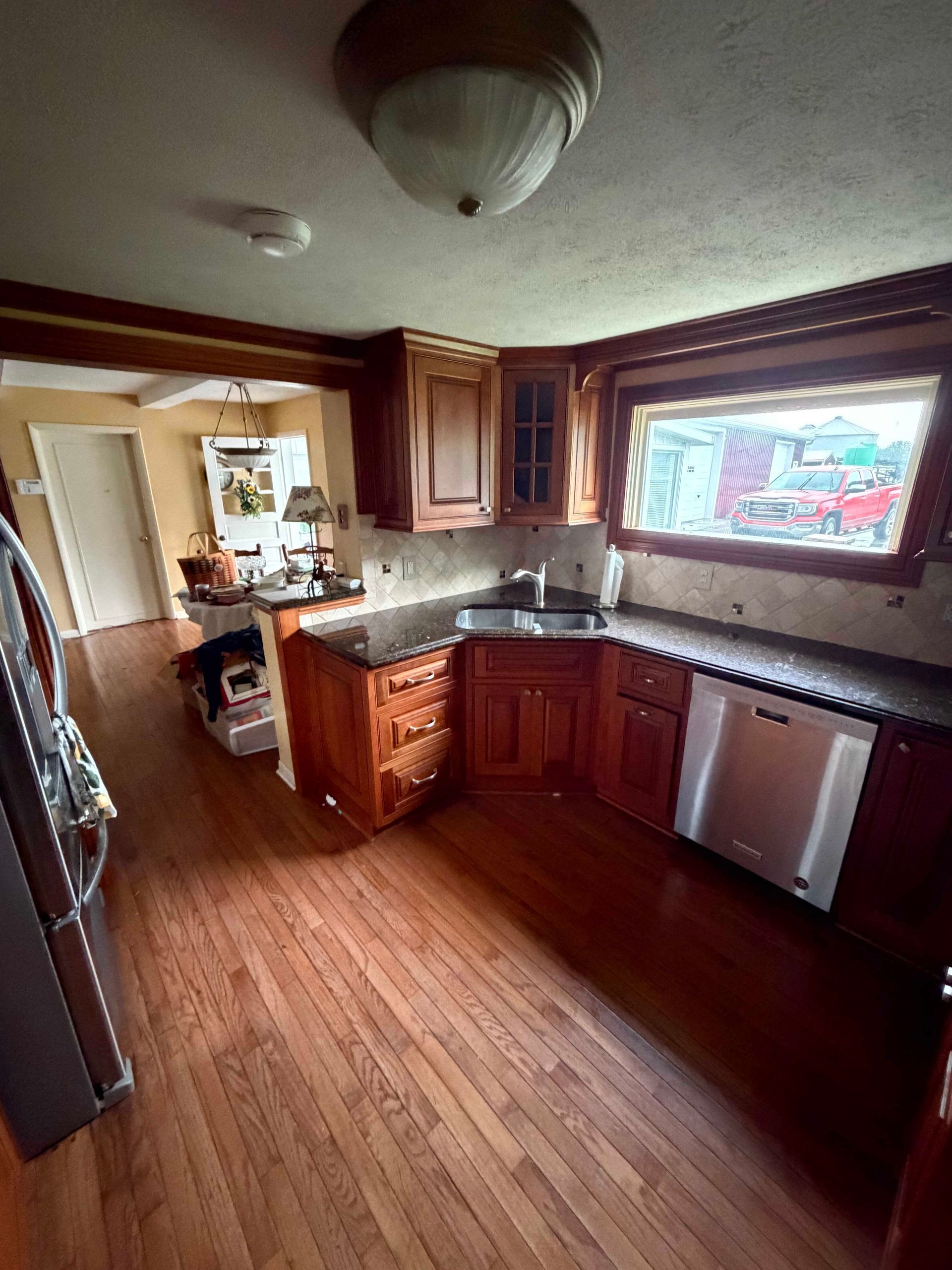 Compact kitchen with wooden cabinets, dark countertops, hardwood floor, and a dishwasher by a bright window.