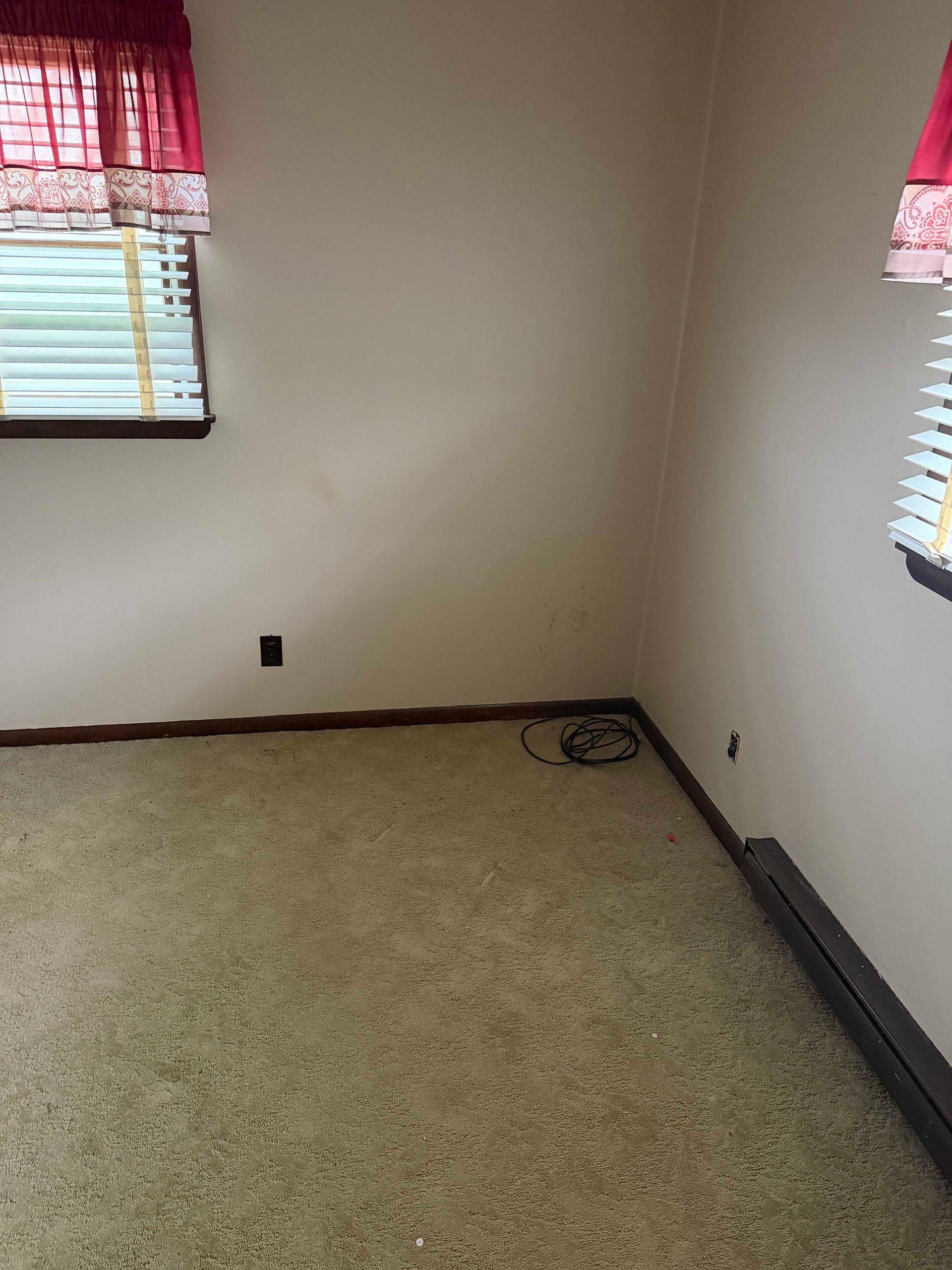 Empty beige room with two windows and red curtains, carpeted floor, and visible cords in the corner