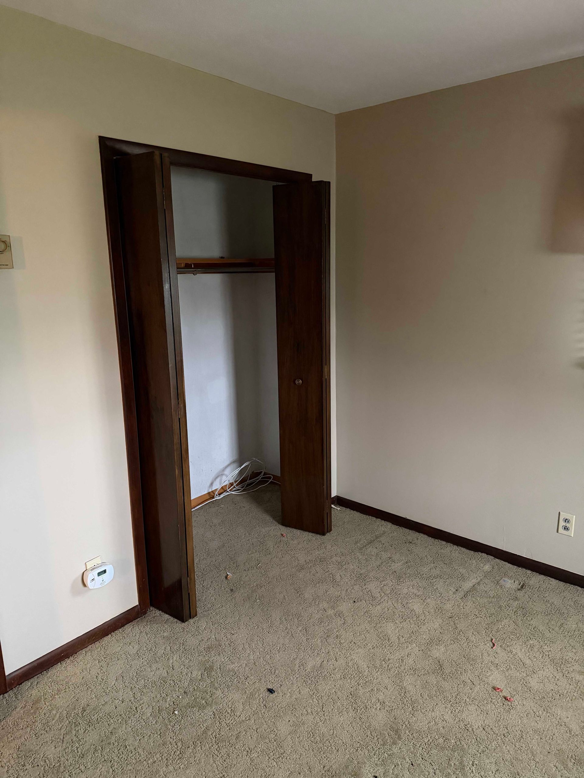 Empty beige bedroom corner with open closet, dark wood trim, and carpeted floor.