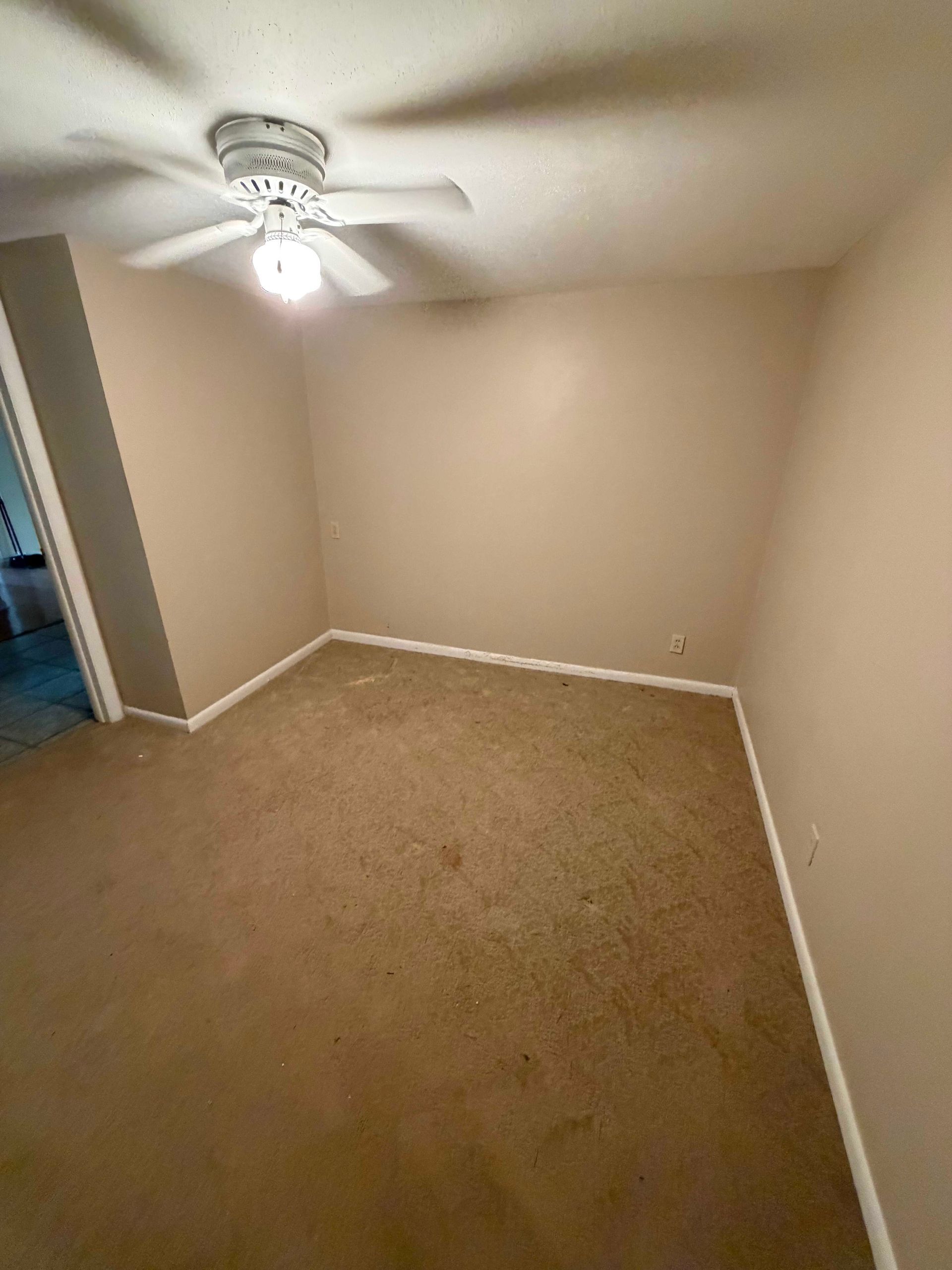 Empty beige bedroom with carpet, white trim, and a ceiling fan/light.