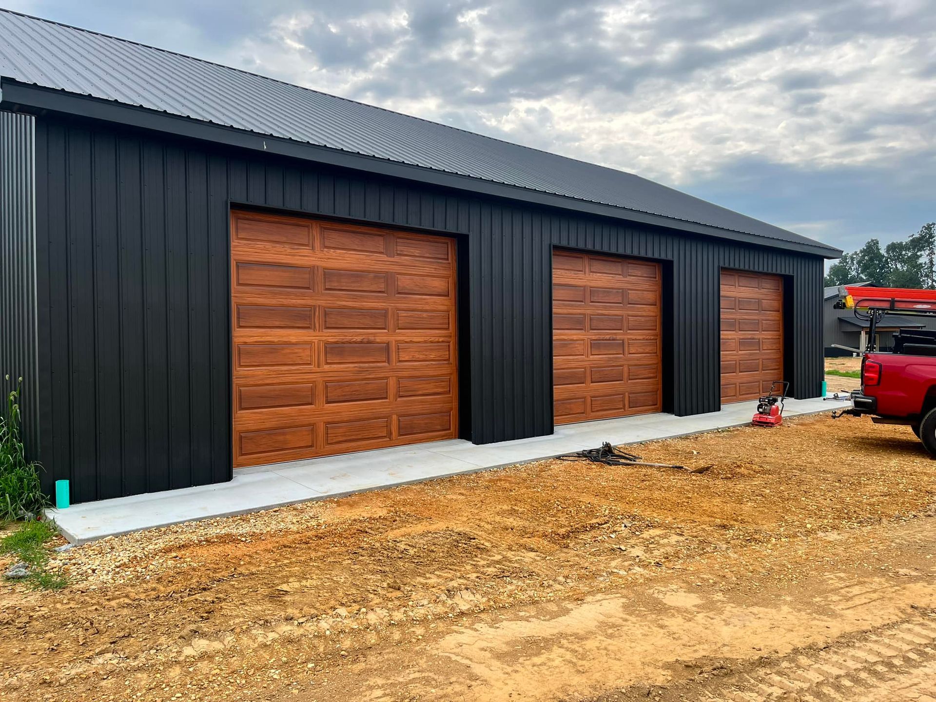 Black building with three brown garage doors on a concrete slab in a dirt lot.