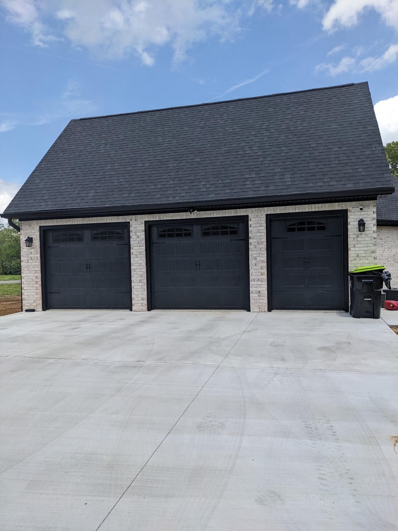Three-car garage with dark gray doors, light brick walls, and a dark shingled roof on a concrete driveway.