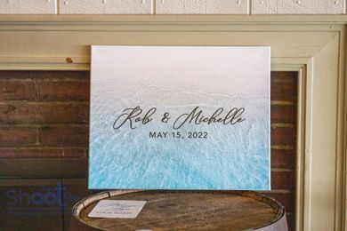 A square canvas on a wooden barrel against a brick fireplace featuring a blue-to-white ombre beach print with text.