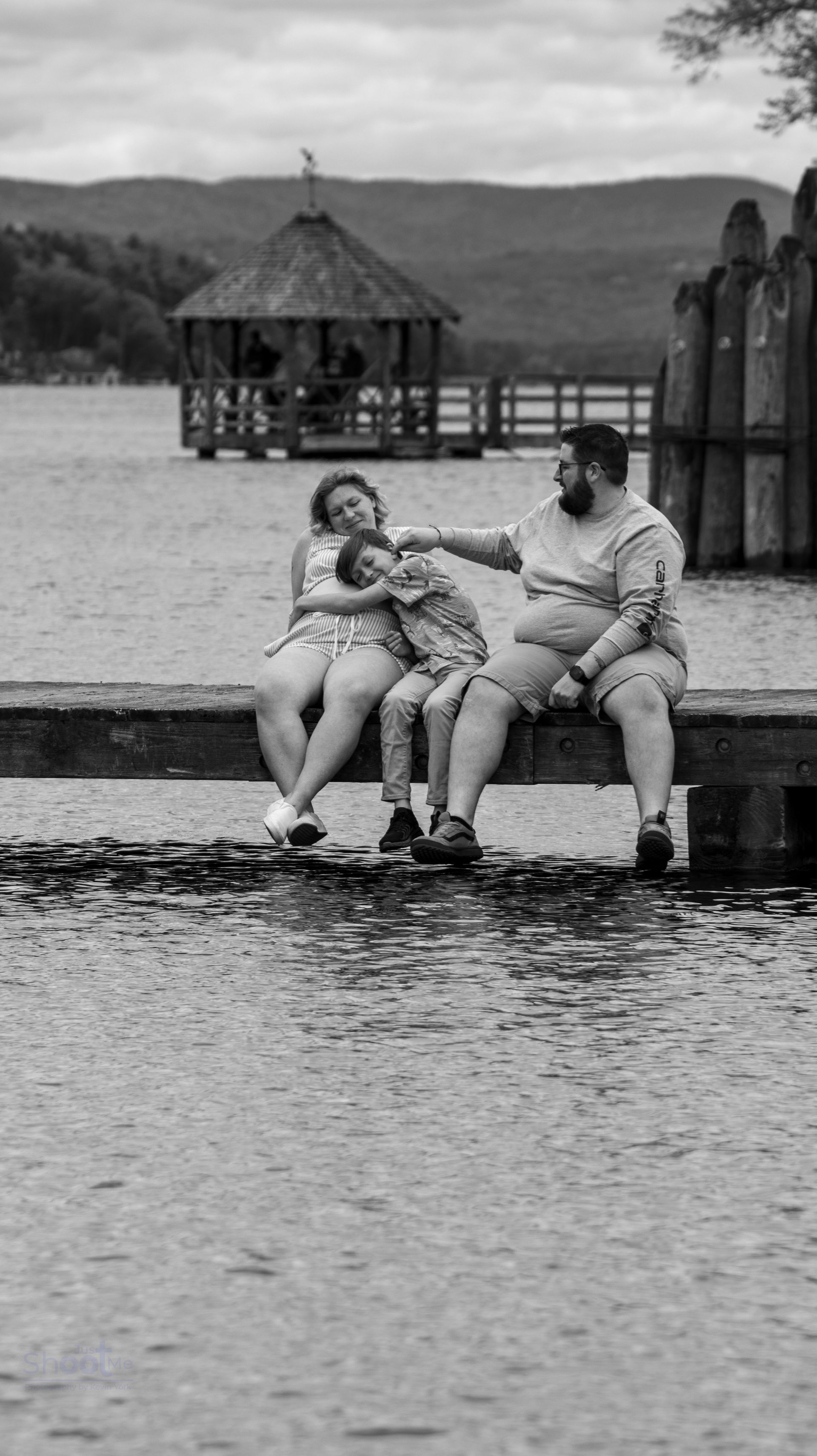 A family of three sits together on a wooden dock overlooking a lake with a gazebo in the background.