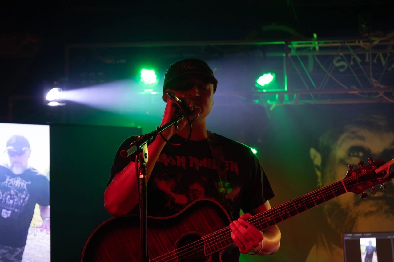 A person in a black cap and t-shirt playing an acoustic guitar and singing into a microphone on a stage with green lights.