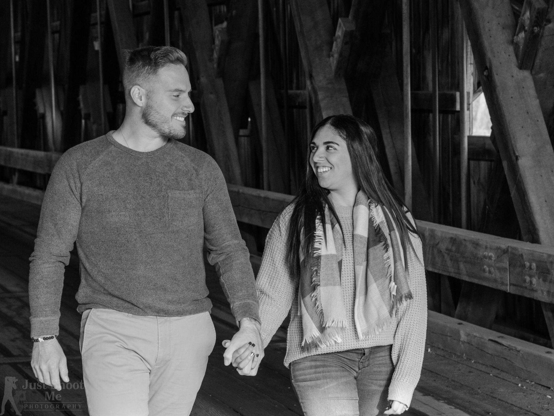 A couple in casual clothing holds hands and smiles while walking through a covered wooden bridge.