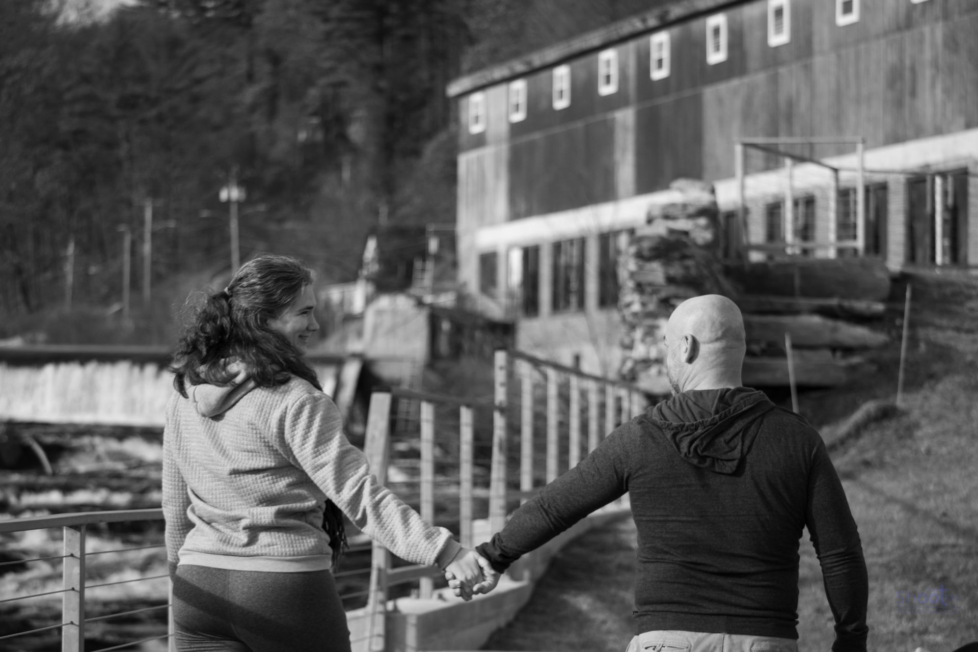A couple holds hands while walking together outdoors near a body of water and an industrial building.