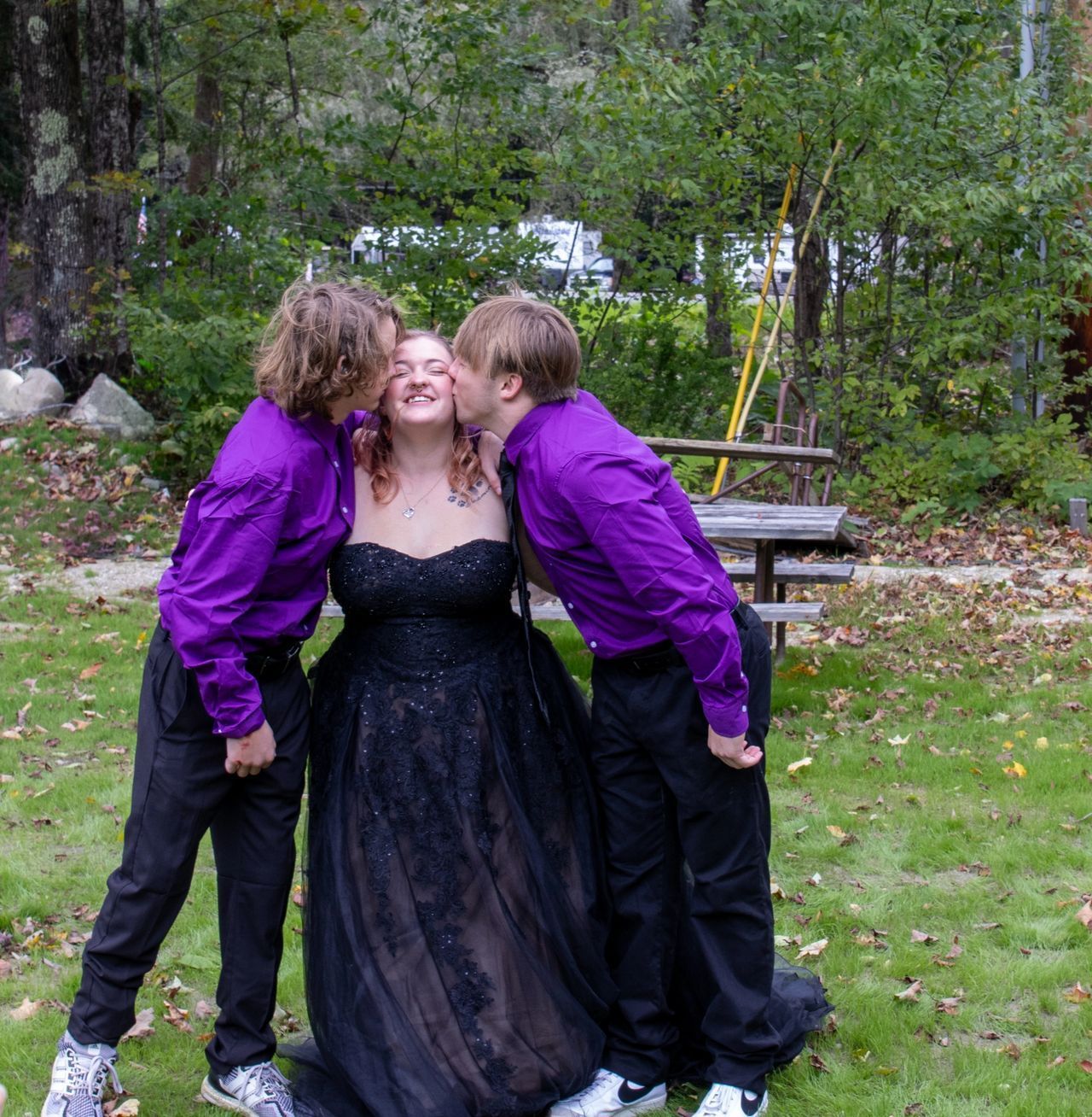 Two people in matching purple shirts kiss a person in a black gown on the cheeks in an outdoor setting.