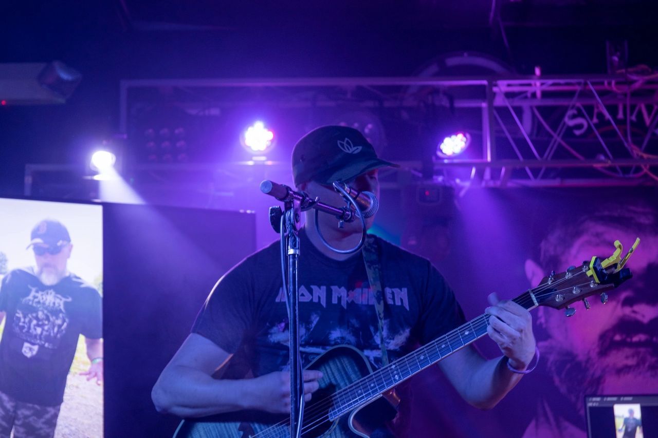 A musician in an Iron Maiden t-shirt and cap plays an acoustic guitar on a stage lit with purple spotlights.
