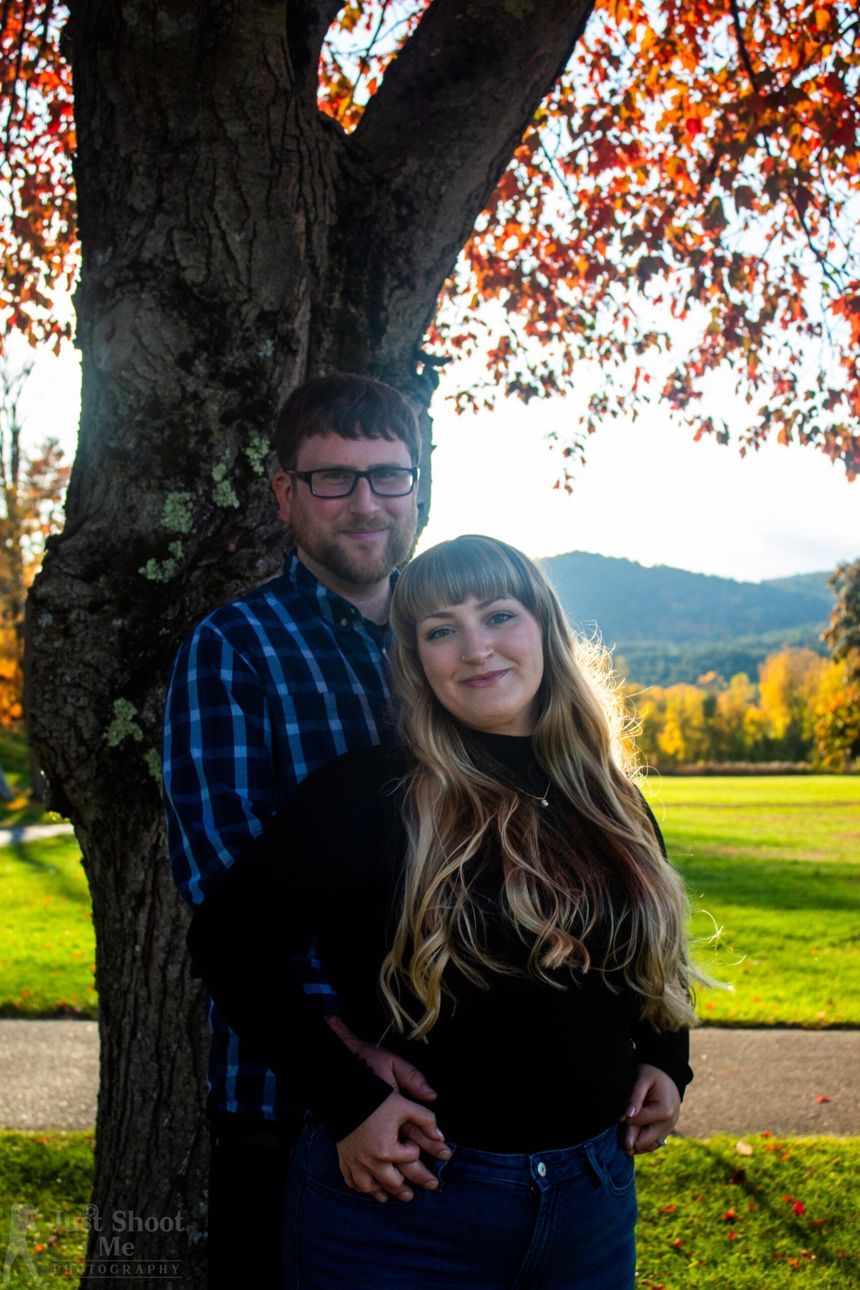 A couple posing under a tree with autumn foliage, mountains in the distance, and a grass field in the background.