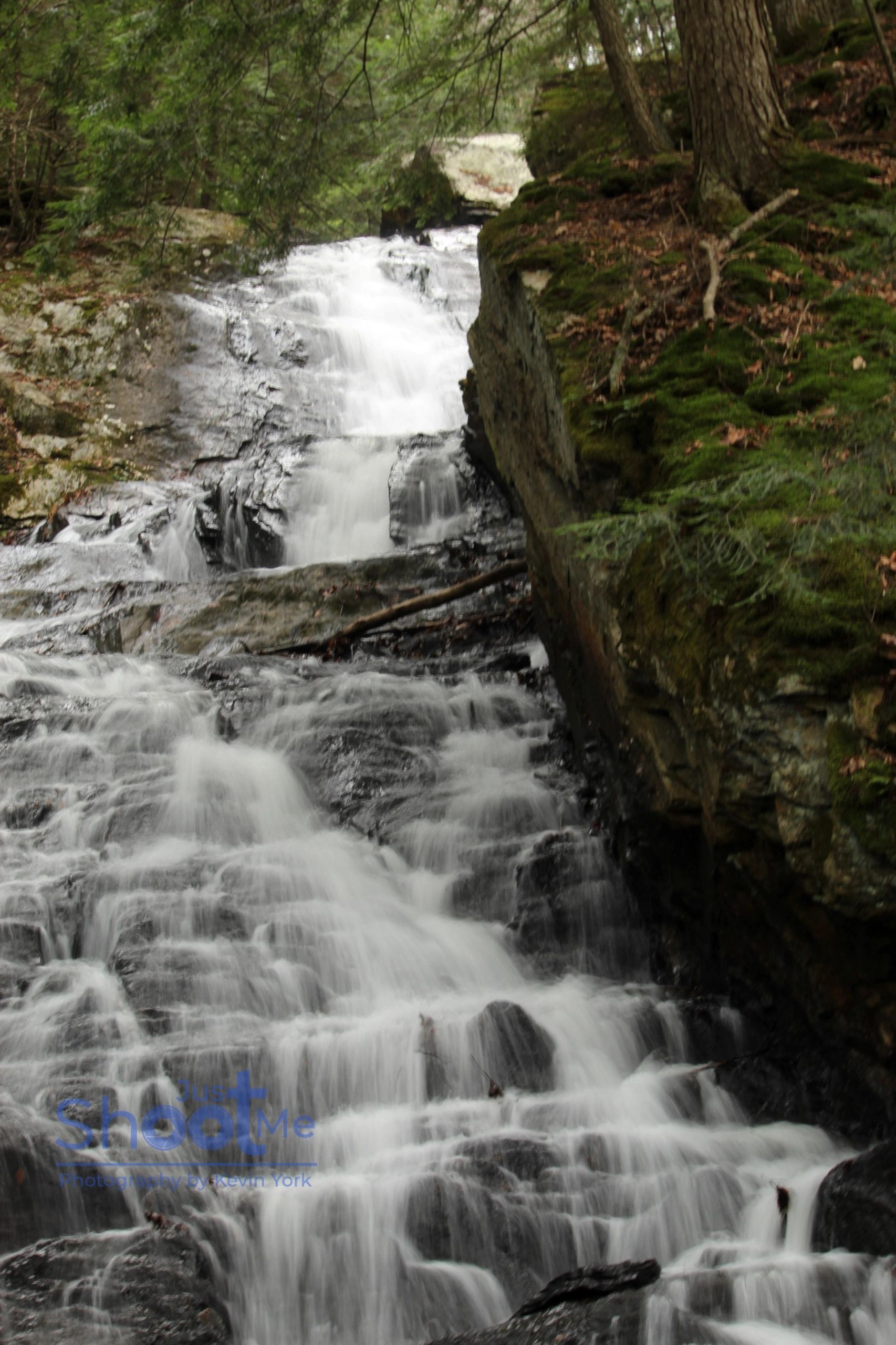 A multi-tiered waterfall cascades down rocky, dark terrain surrounded by lush green forest foliage.