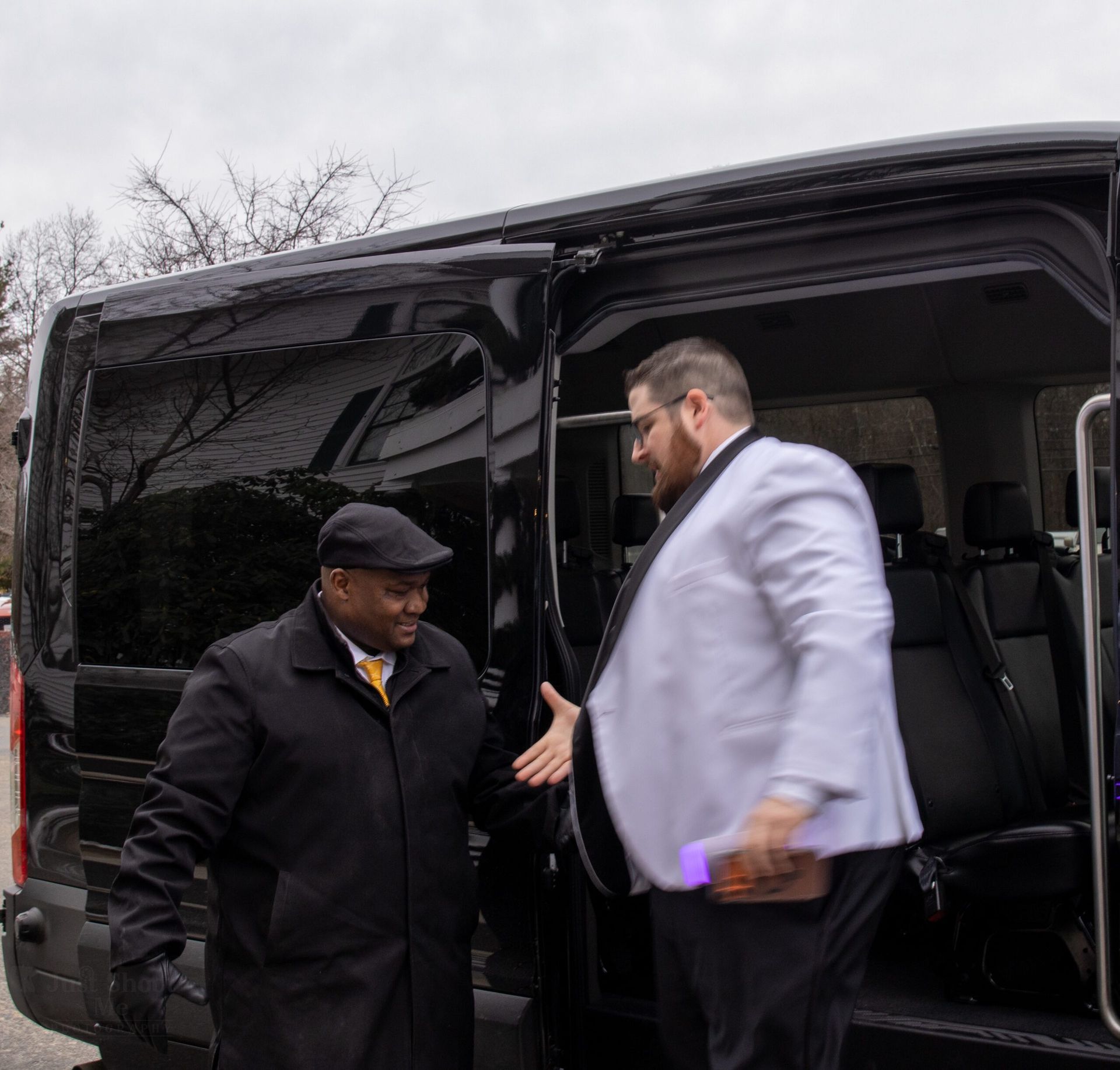 Two men, one in a dark coat and cap and the other in a light jacket, exchange a handshake outside a black van.