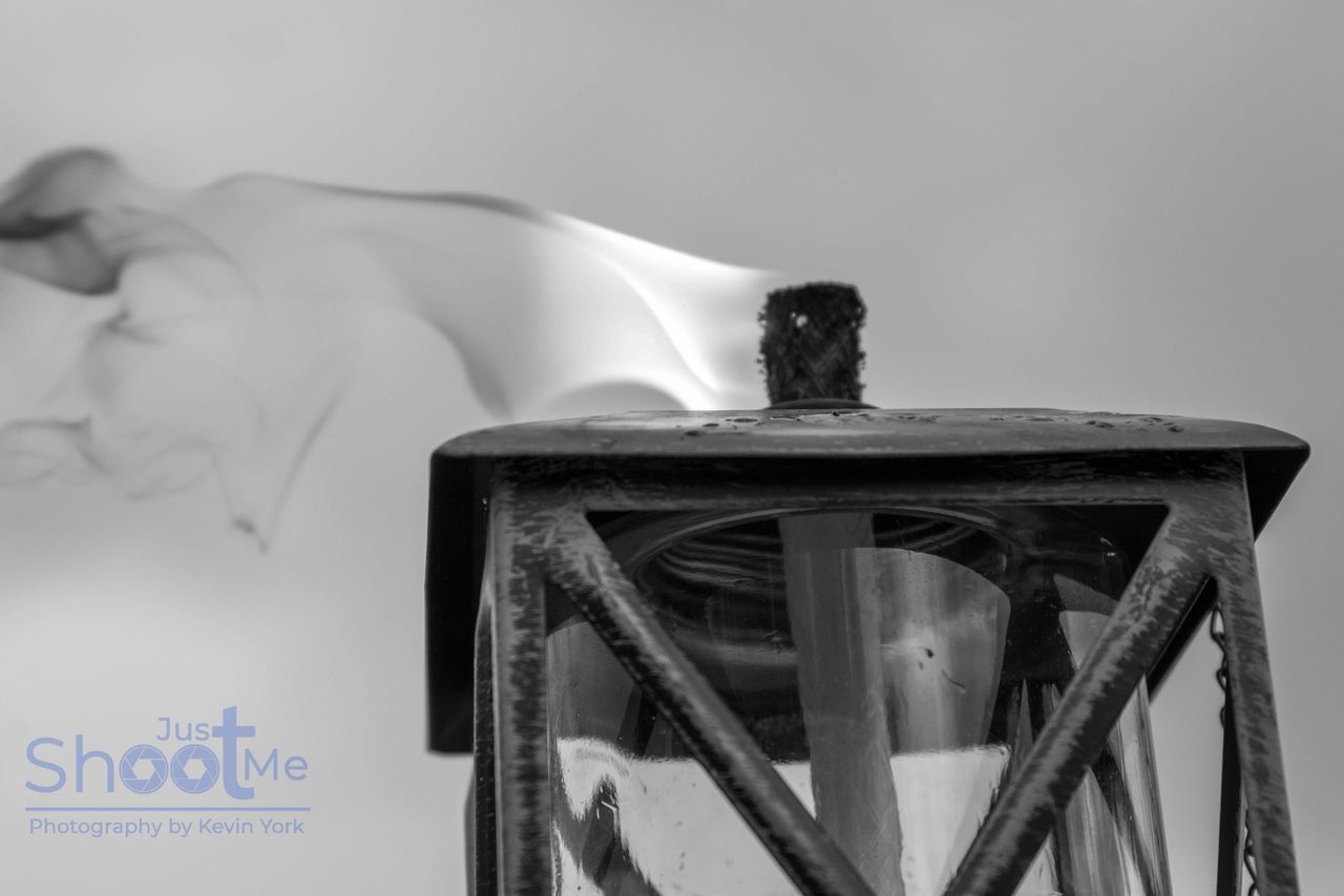 Black and white photo of a dark metal lantern with a wisp of smoke rising from its top against a plain background.