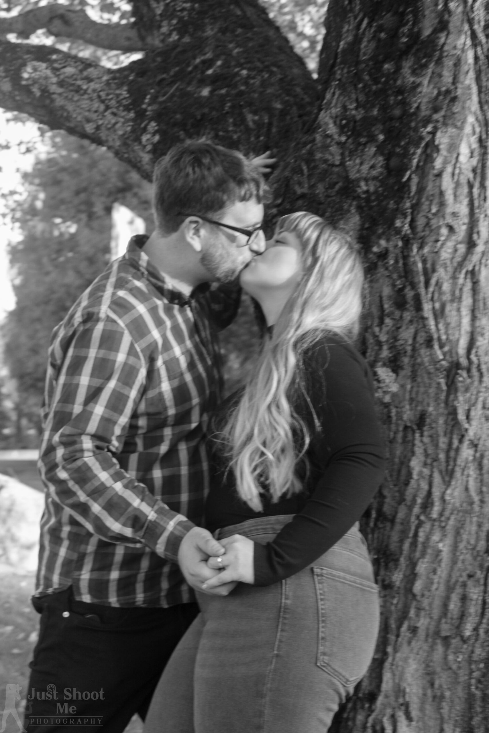 A black and white photo of a couple kissing while standing hand-in-hand next to a large tree.