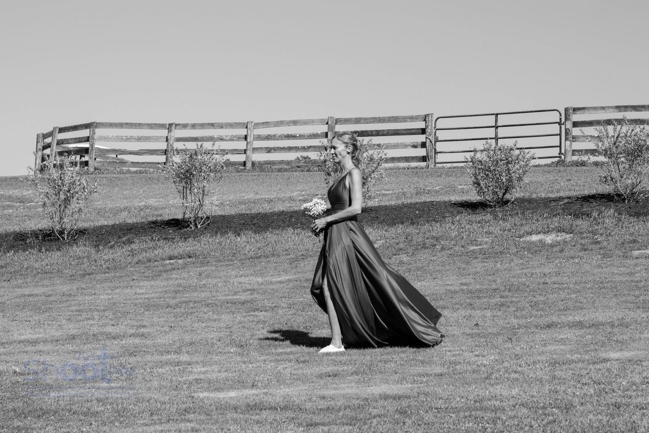 A person in a long, flowing gown with a high slit walks across a field toward a wooden fence.