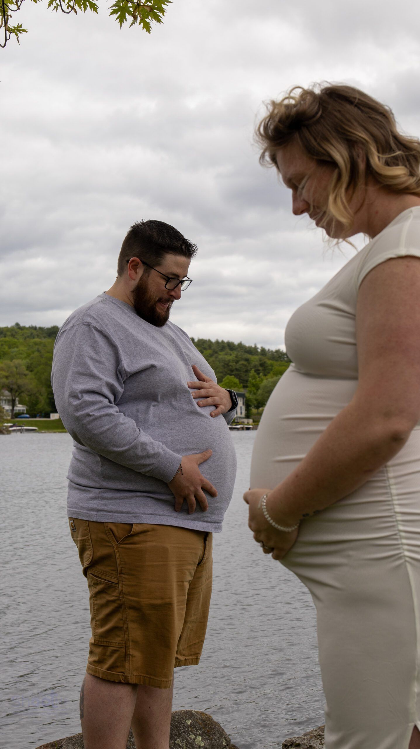 A person and a pregnant individual stand by a lake, both holding their bellies, with trees in the background.
